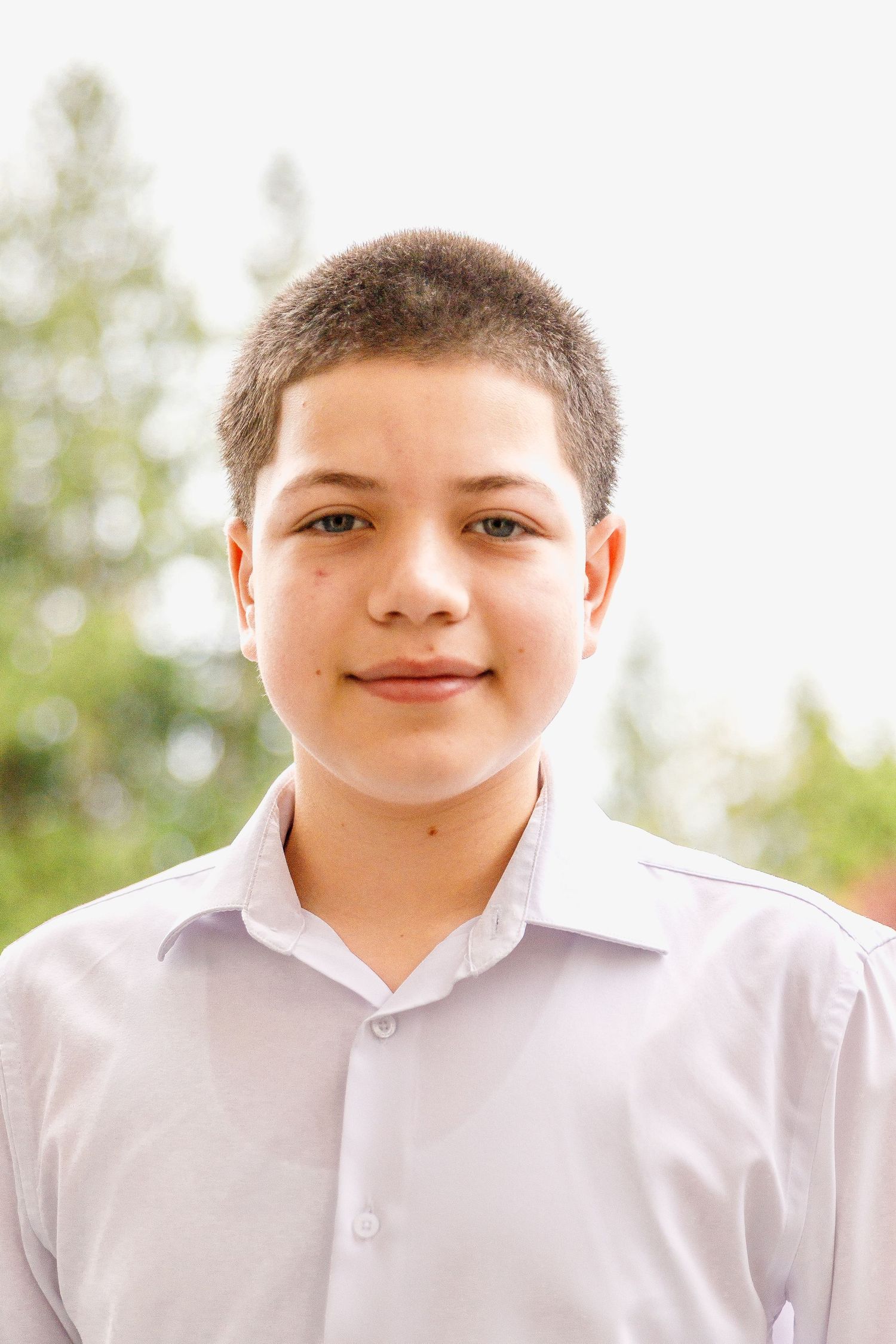 A young person in a white collared shirt smiles pleasantly against an outdoor background with trees.
