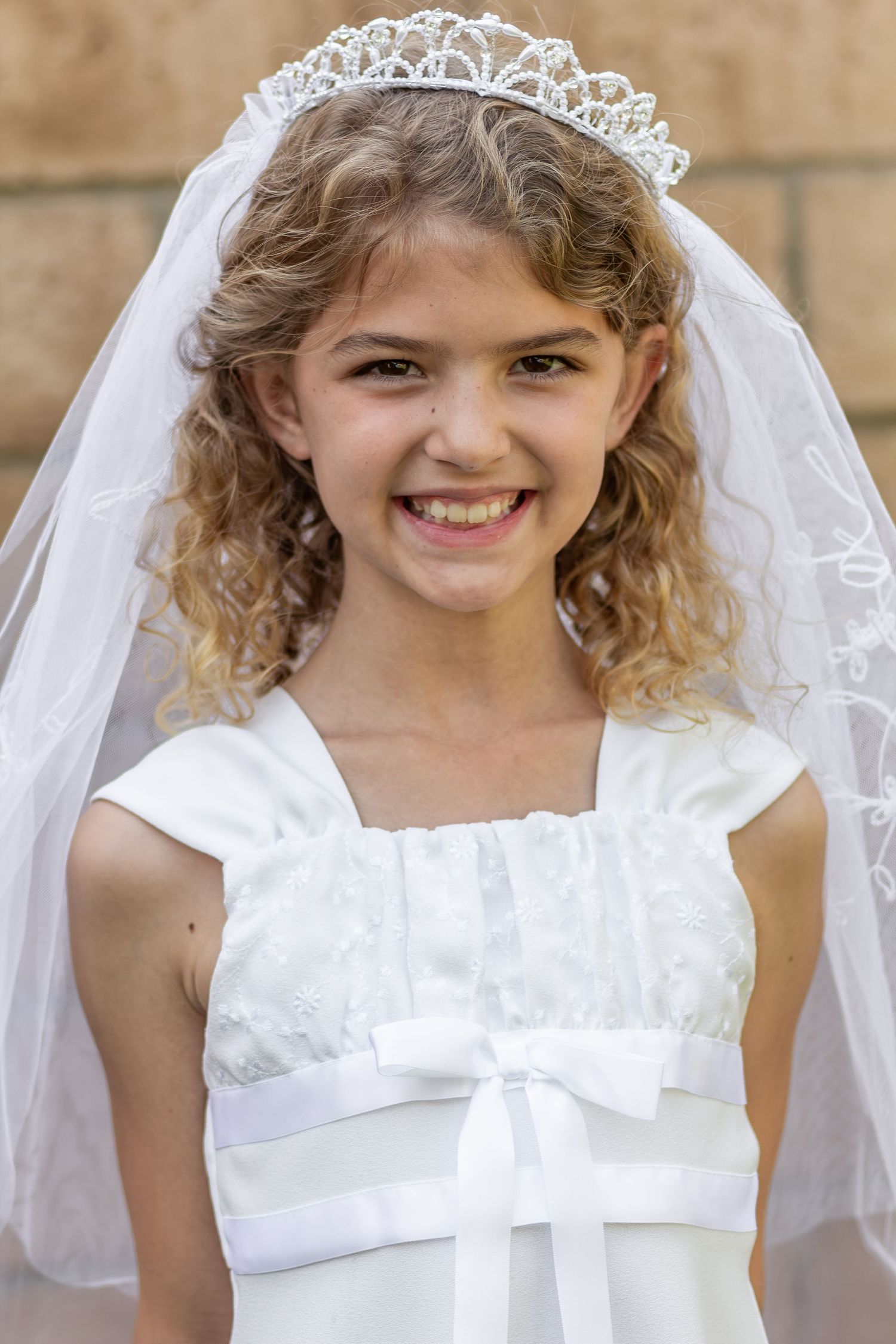 A young person in a white First Communion dress with a lace veil and tiara smiles in an outdoor portrait.