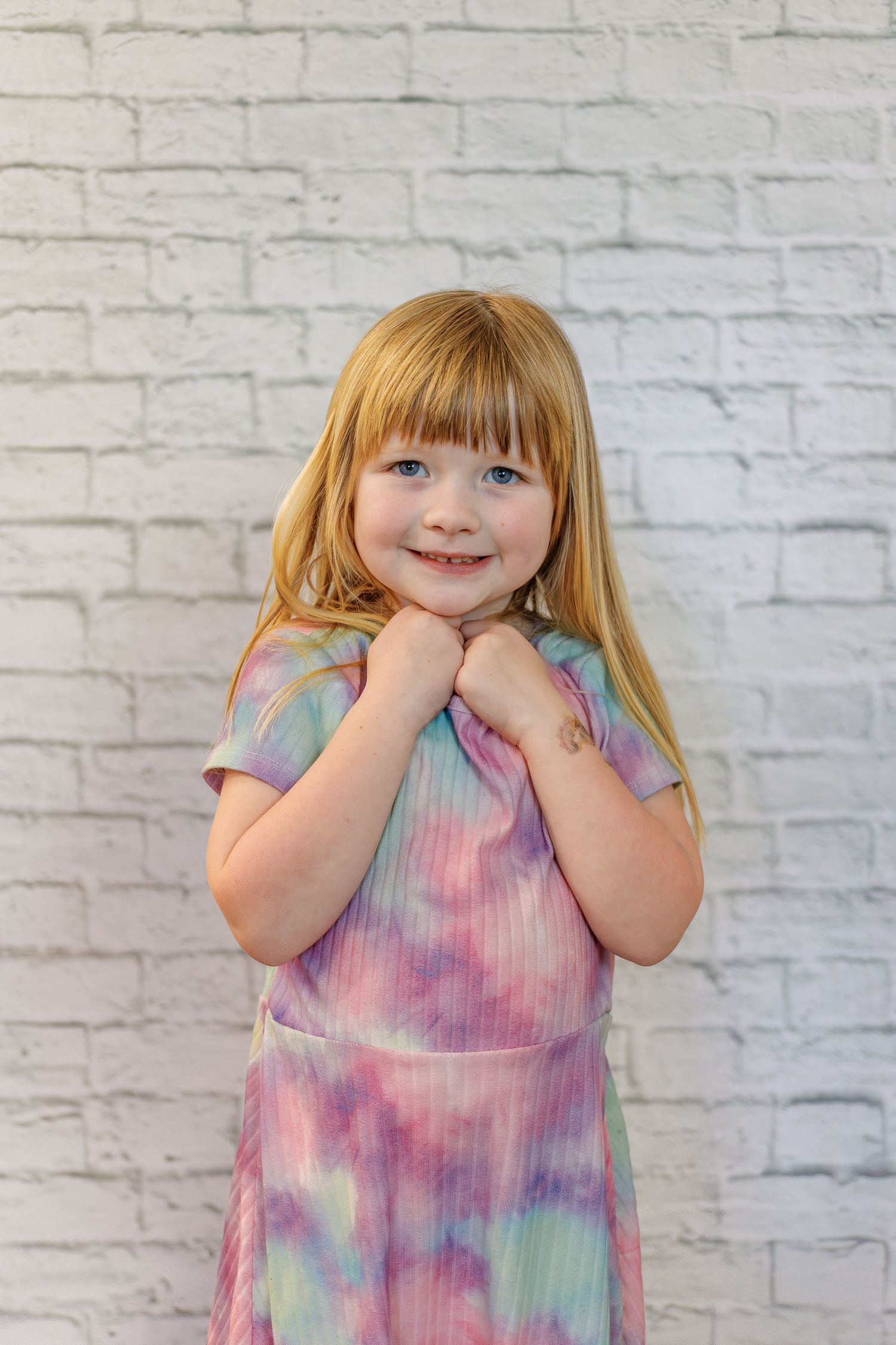 Young child in tie dye pastel dress poses against white brick wall with hands clasped under chin.