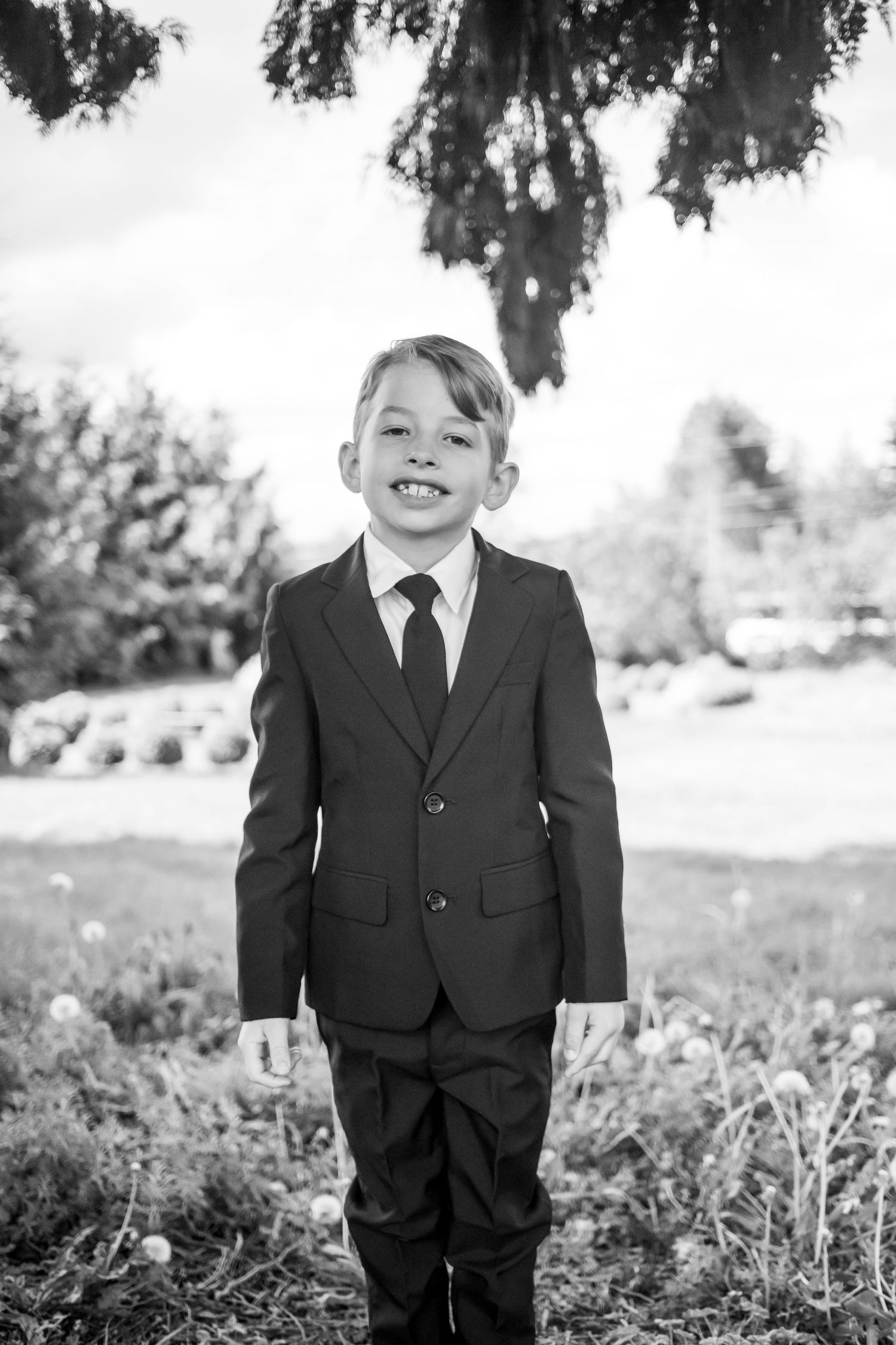 Young child dressed in a formal black suit and tie stands outdoors smiling in a black and white portrait.