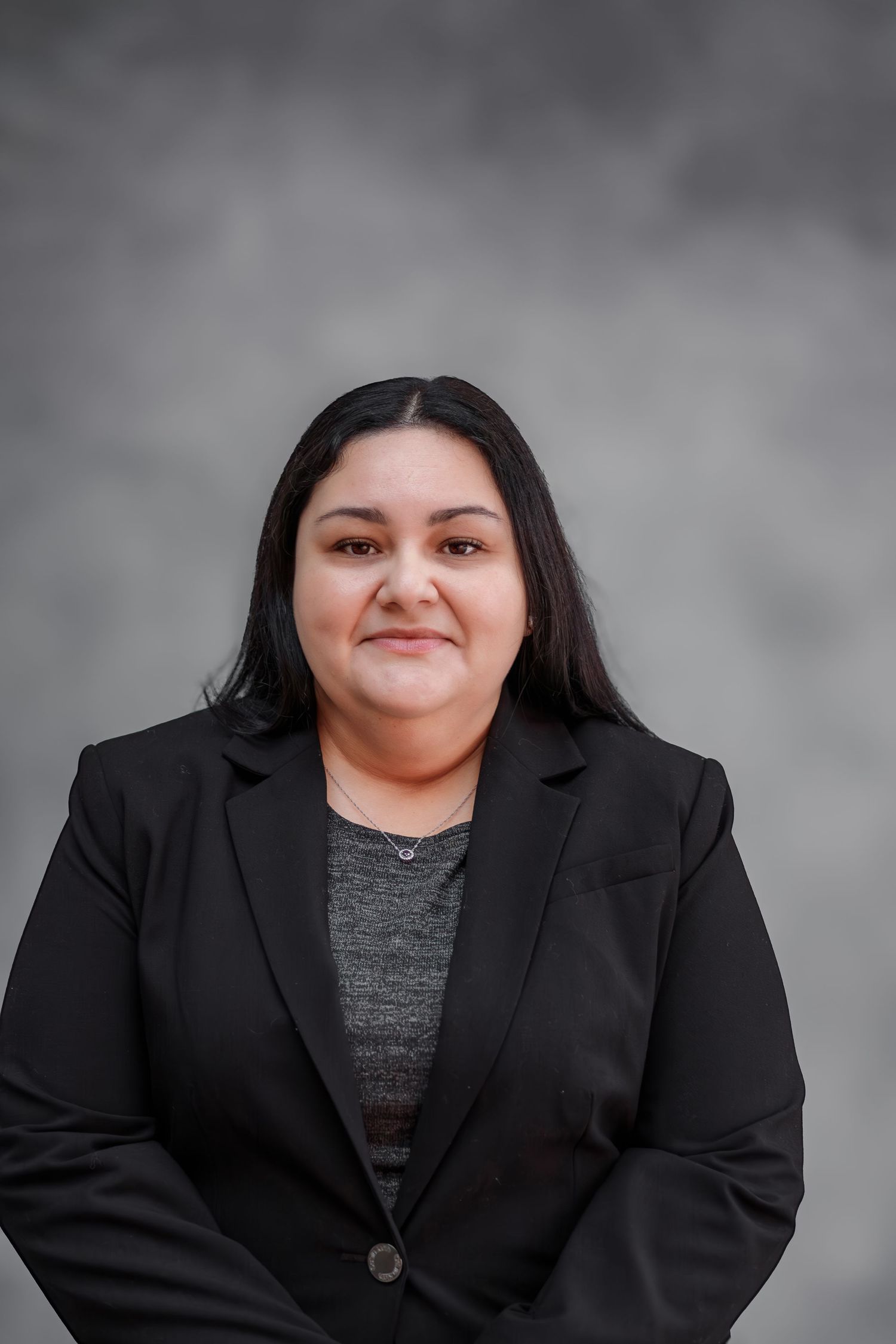 Professional headshot of a young woman with black hair wearing a black blazer against a gray background.