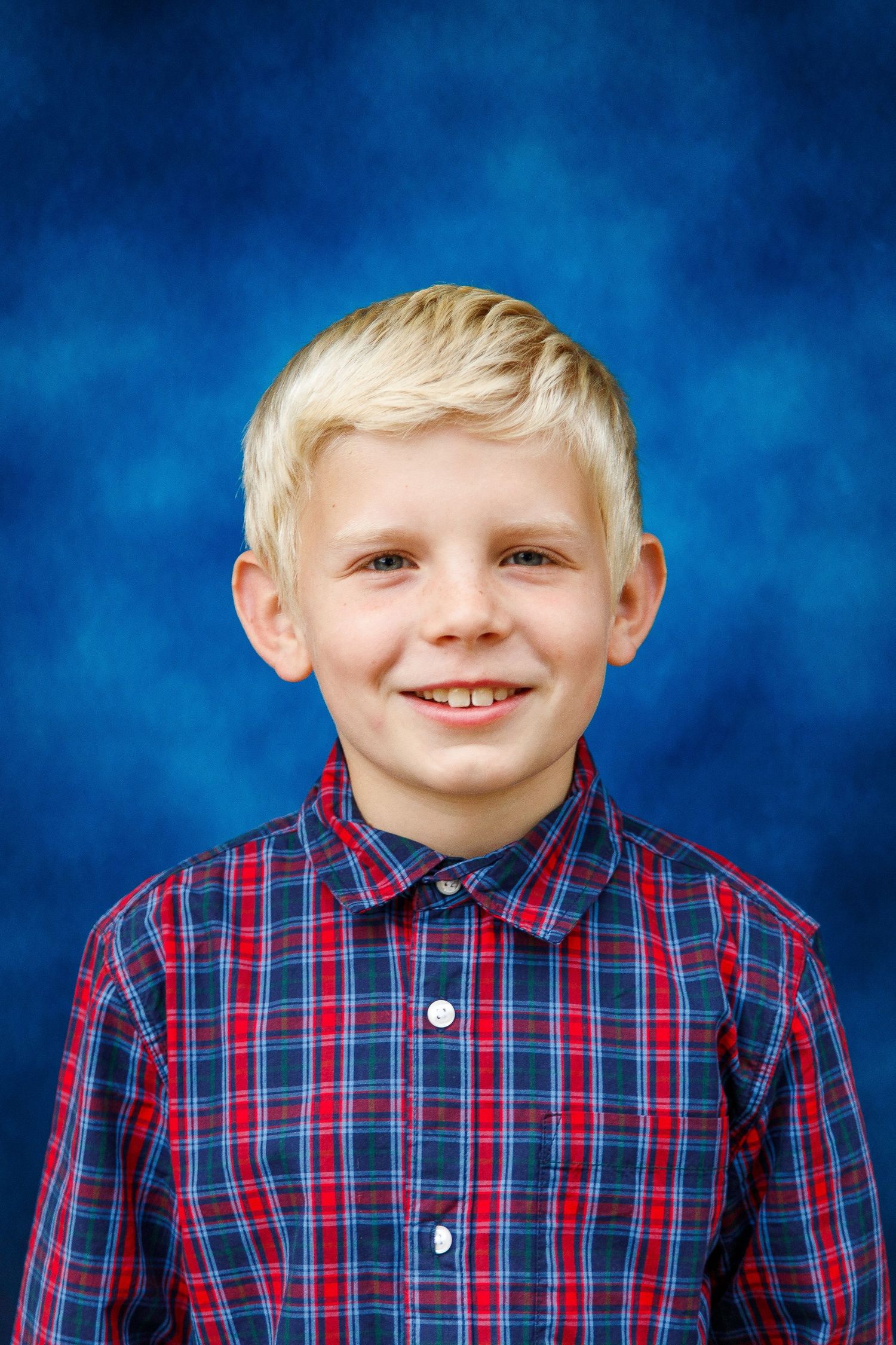 Young student in red and blue plaid button-up shirt poses for portrait against blue backdrop.