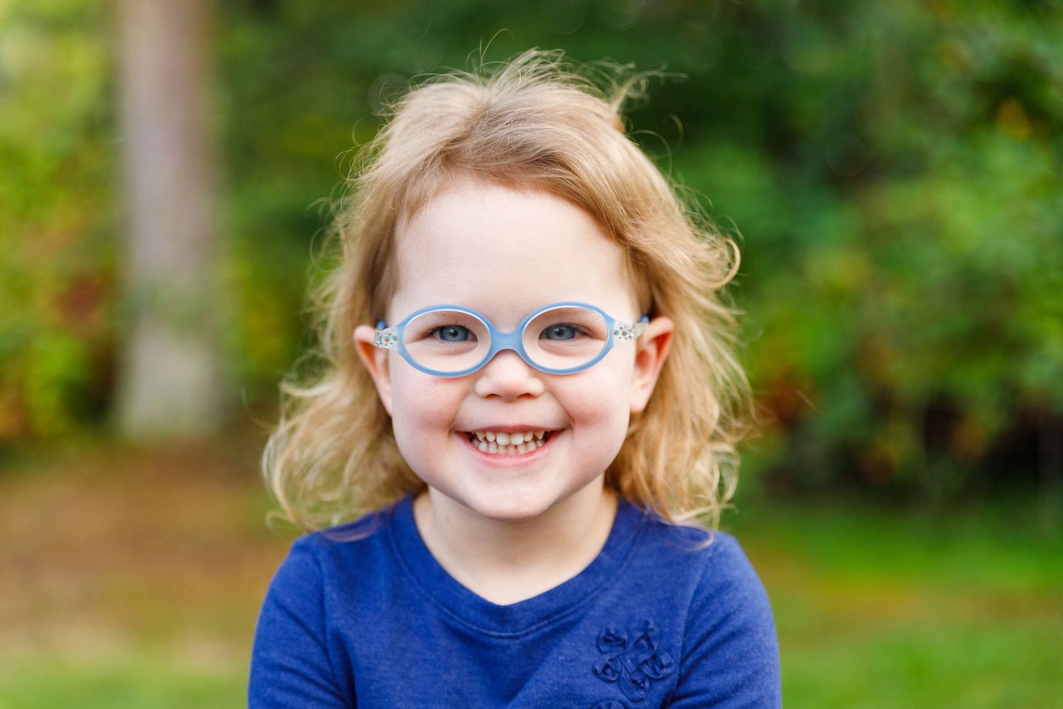 Child in blue glasses and shirt poses with varying expressions outdoors in a green garden setting.