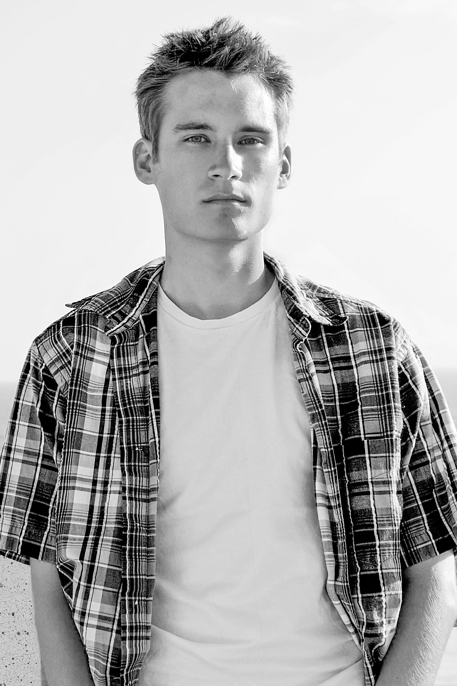 Black and white portrait photo of someone wearing a plaid button-up shirt over a white t-shirt against a light background.