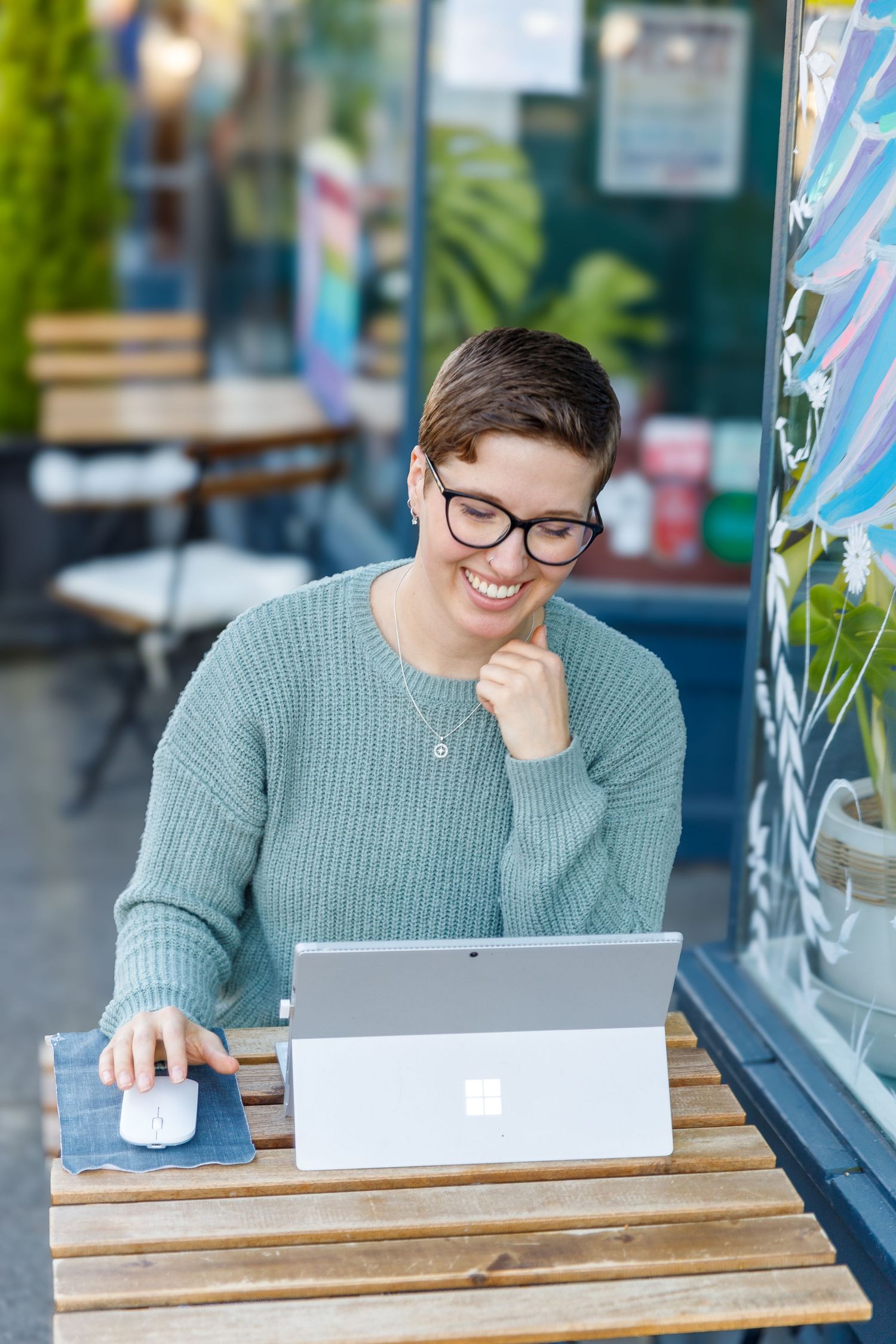 Person in glasses and gray sweater sits at wooden table with laptop and documents in bright cafe setting.