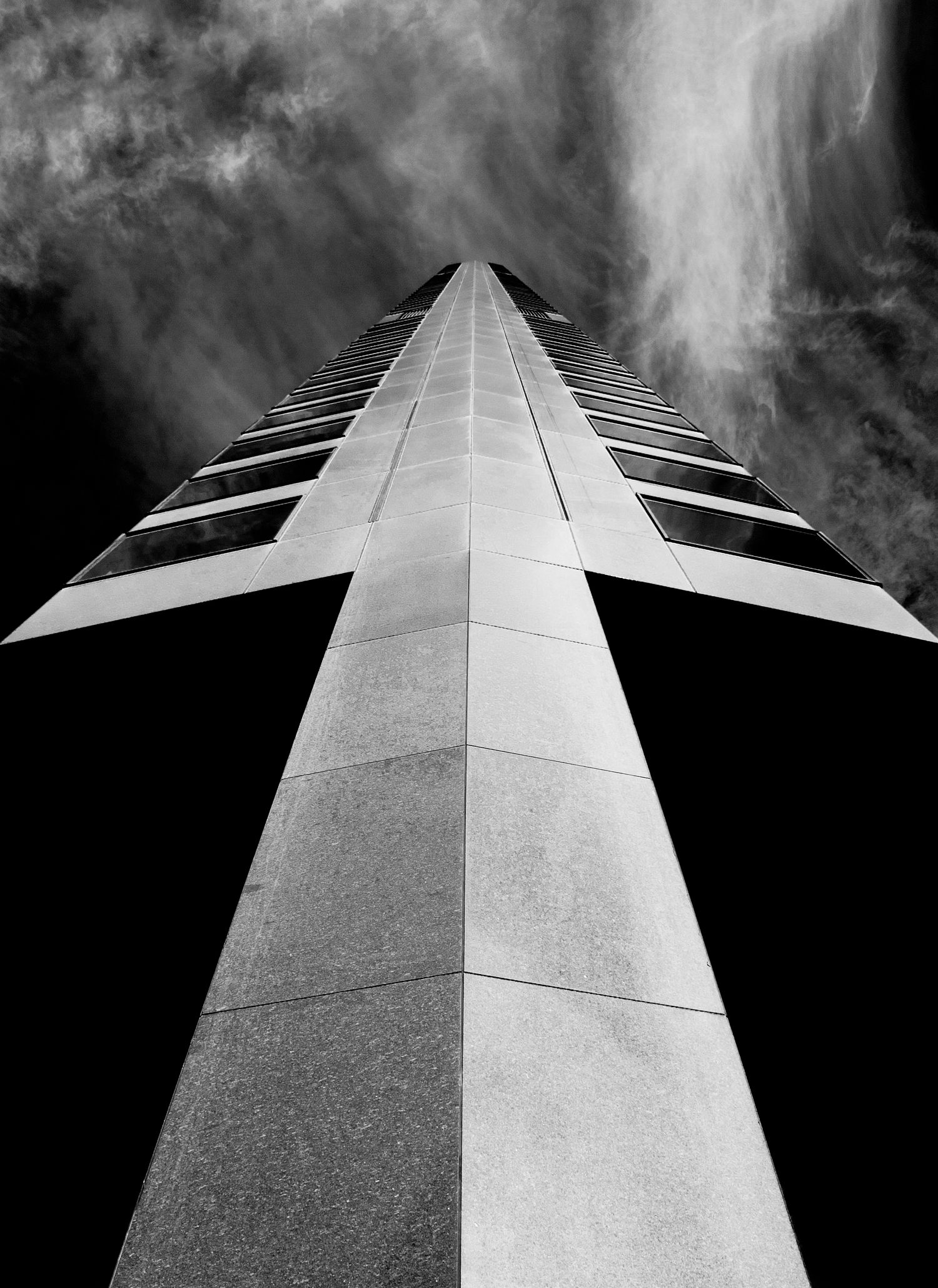 Dramatic upward view of a modern skyscraper against cloudy sky in black and white.