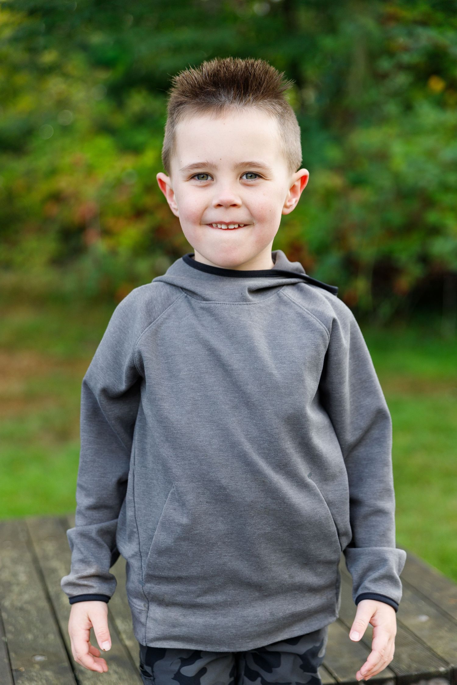 Child in a cozy gray sweatshirt poses outdoors on a wooden bench with greenery in the background.