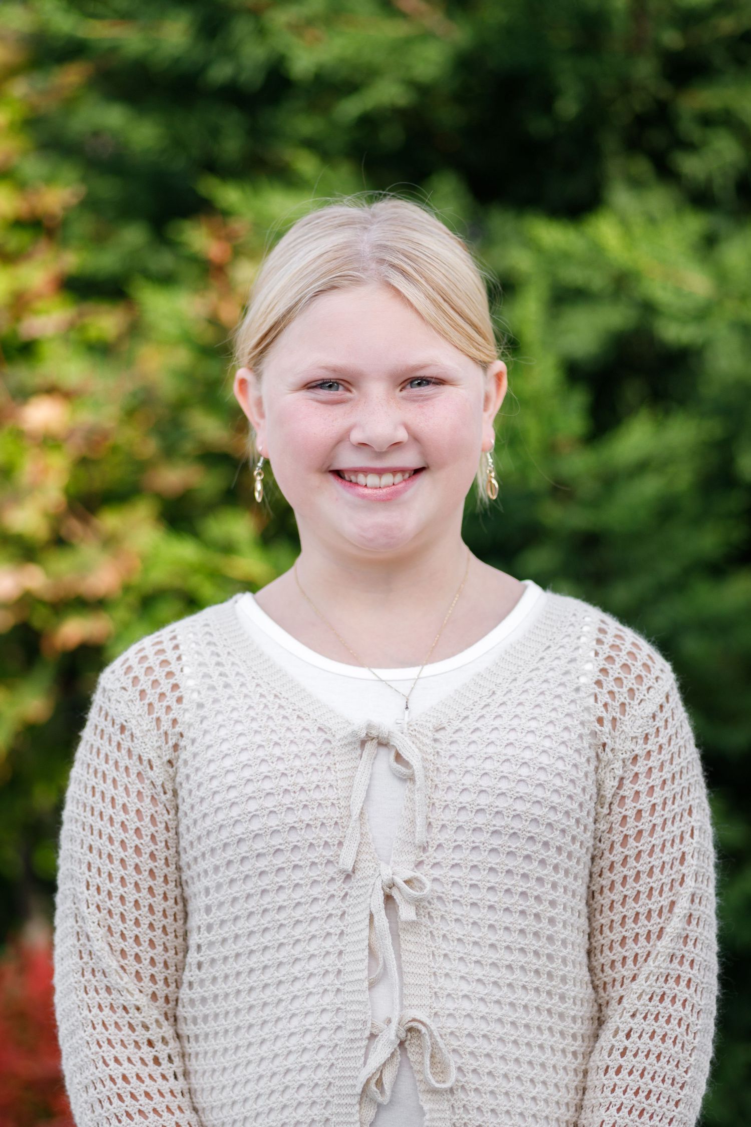 A youth in a cream-colored knit sweater with tie detail smiles while standing outdoors.