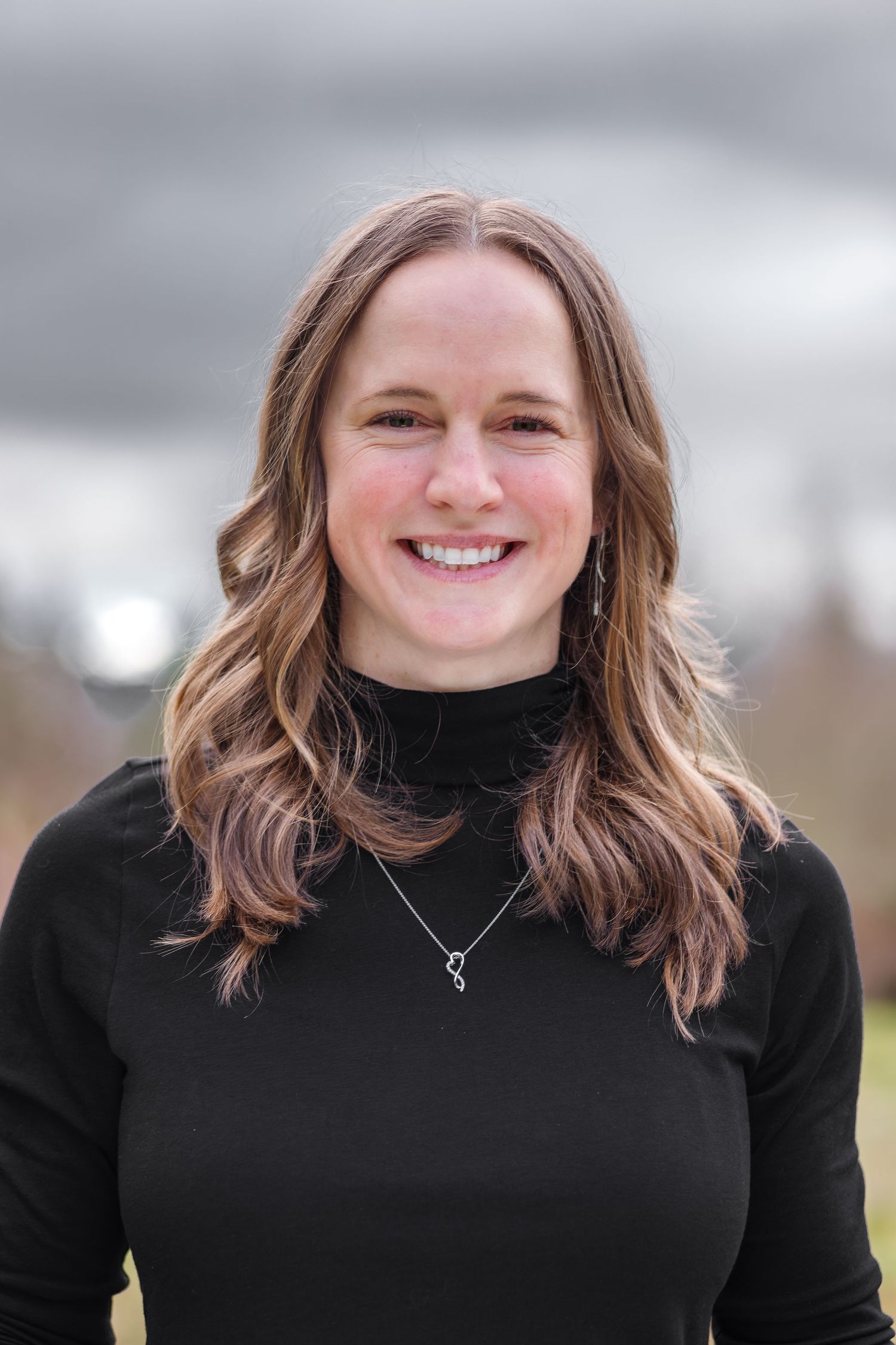 Smiling young woman with long brown hair wearing a black turtleneck and silver necklace outdoors.