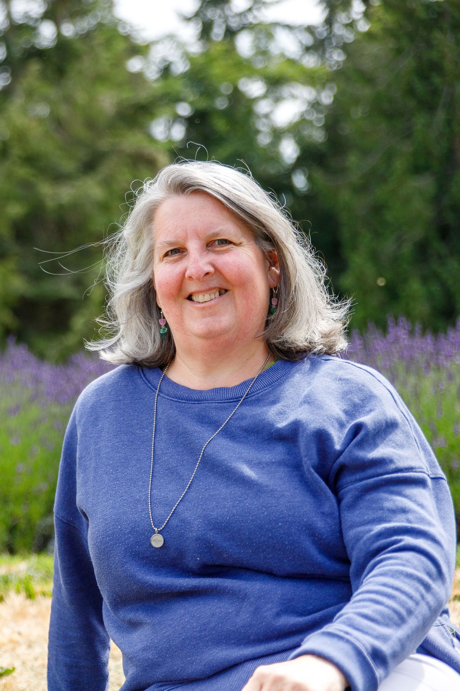 Someone wearing a blue sweater and necklace sits outdoors with purple wildflowers blooming in the background.
