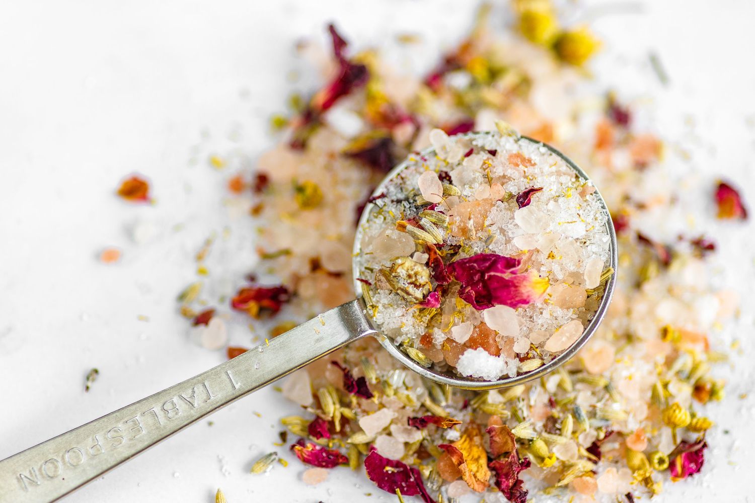 Dried floral bath salts with rose petals and herbs displayed on a vintage measuring spoon against white background.