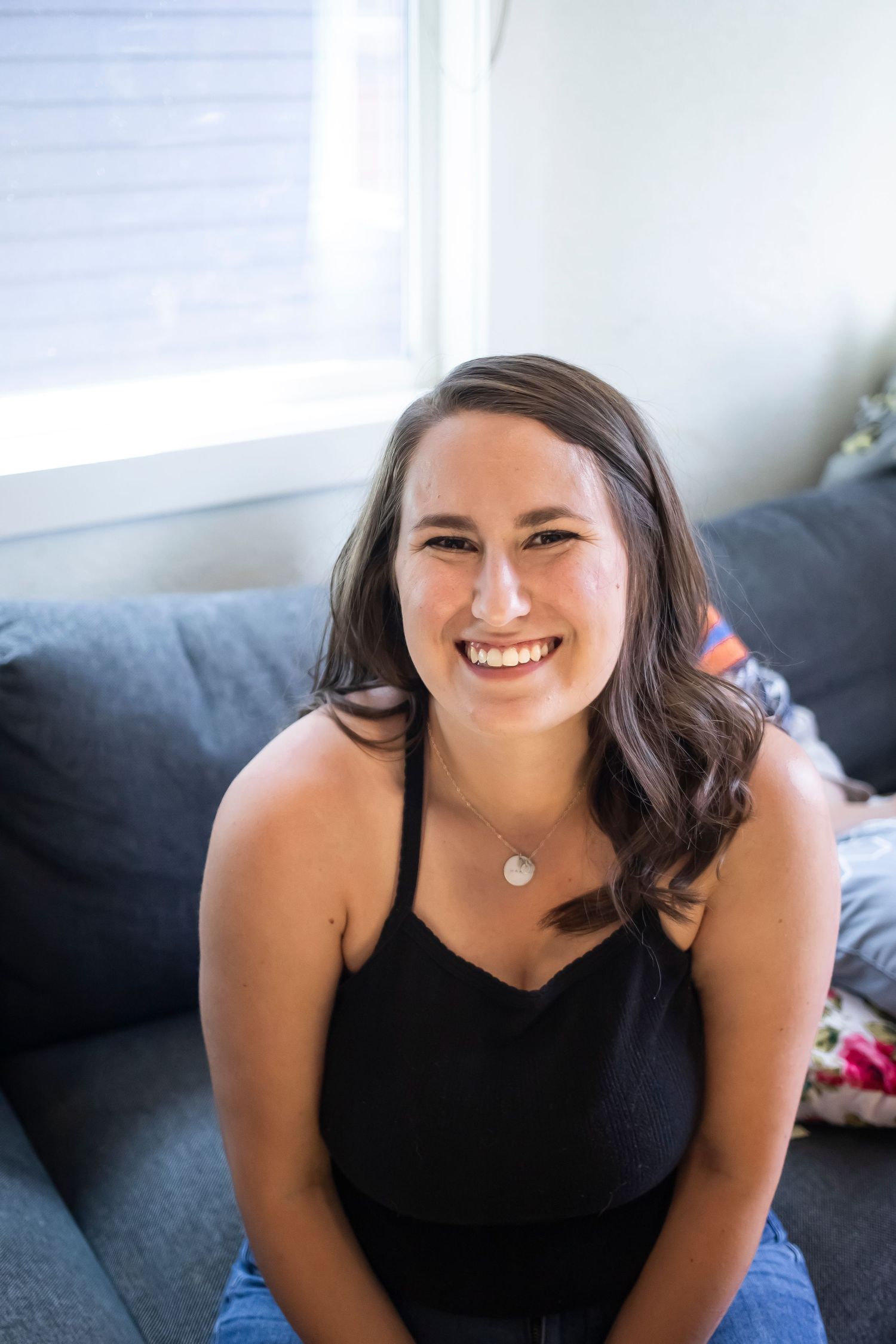 Relaxed indoor portrait of woman in black top sitting on grey couch.