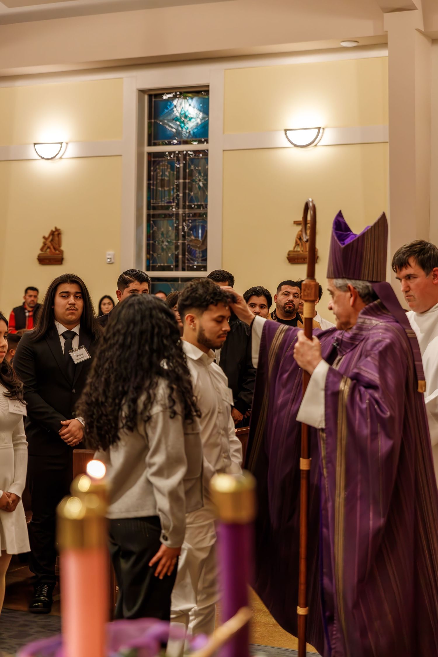 Religious ceremony in a church showing attendees gathered around a bishop in purple robes near a stained glass window.