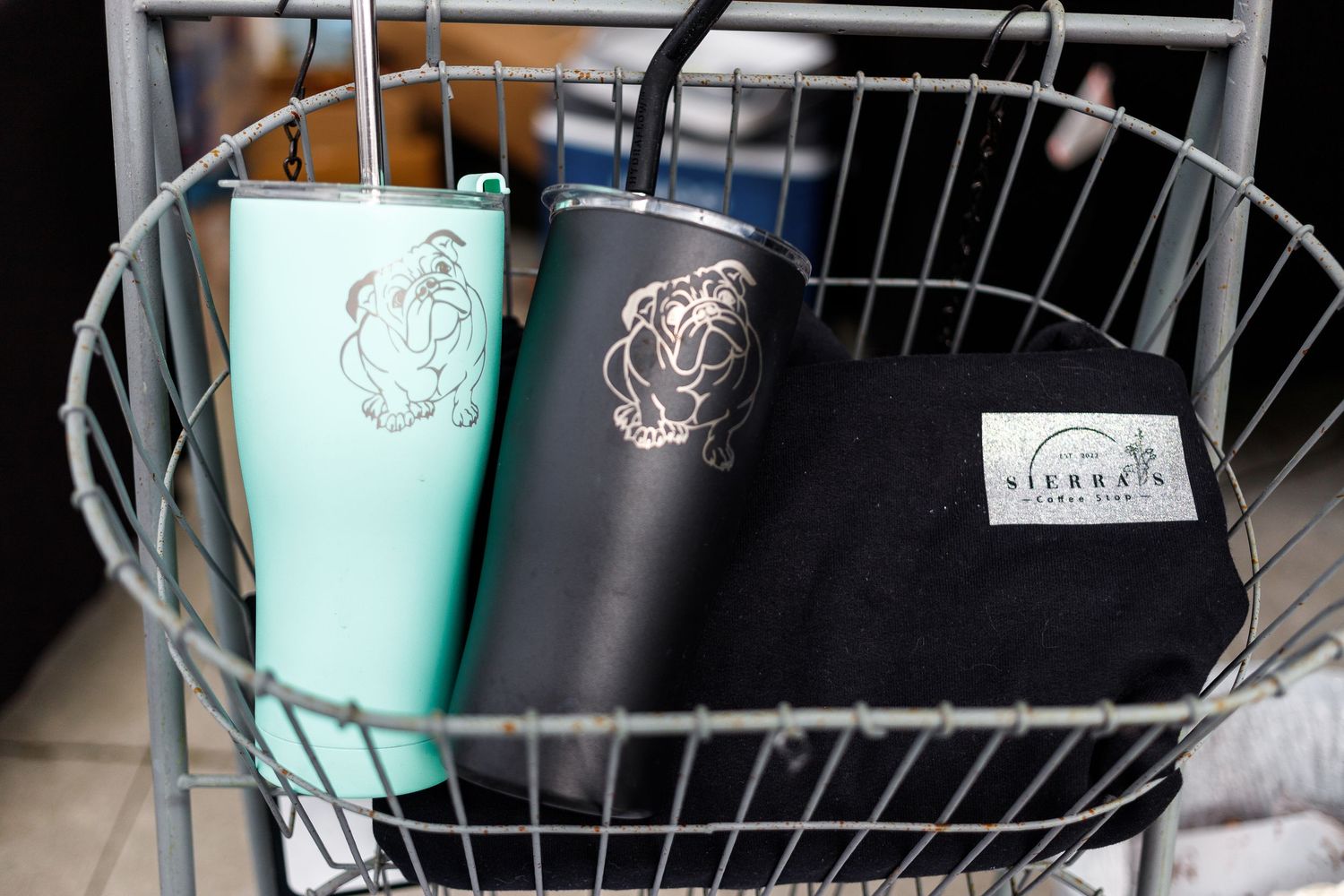 Two thermal tumblers in mint green and black with monogram designs sit in a wire basket with a planner.