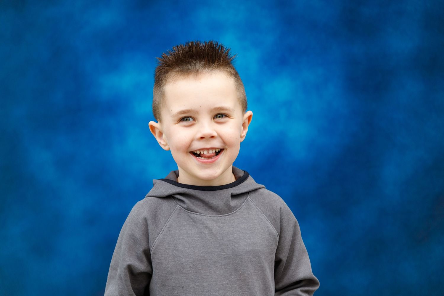 Child wearing a gray hoodie smiles brightly against a mottled blue background in a studio portrait.