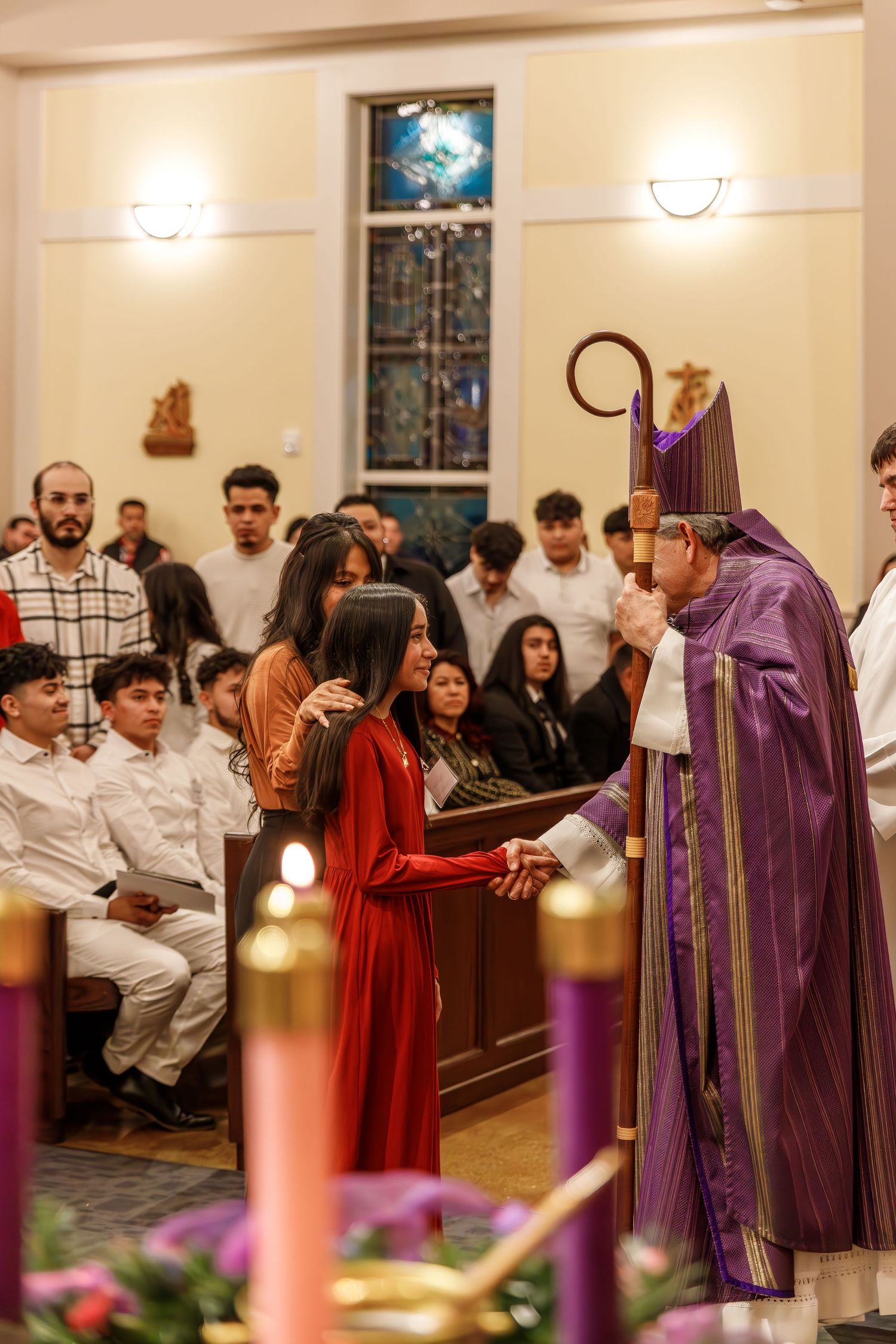 A religious ceremony with purple-robed clergy and congregation gathered in a church with candles and stained glass windows.