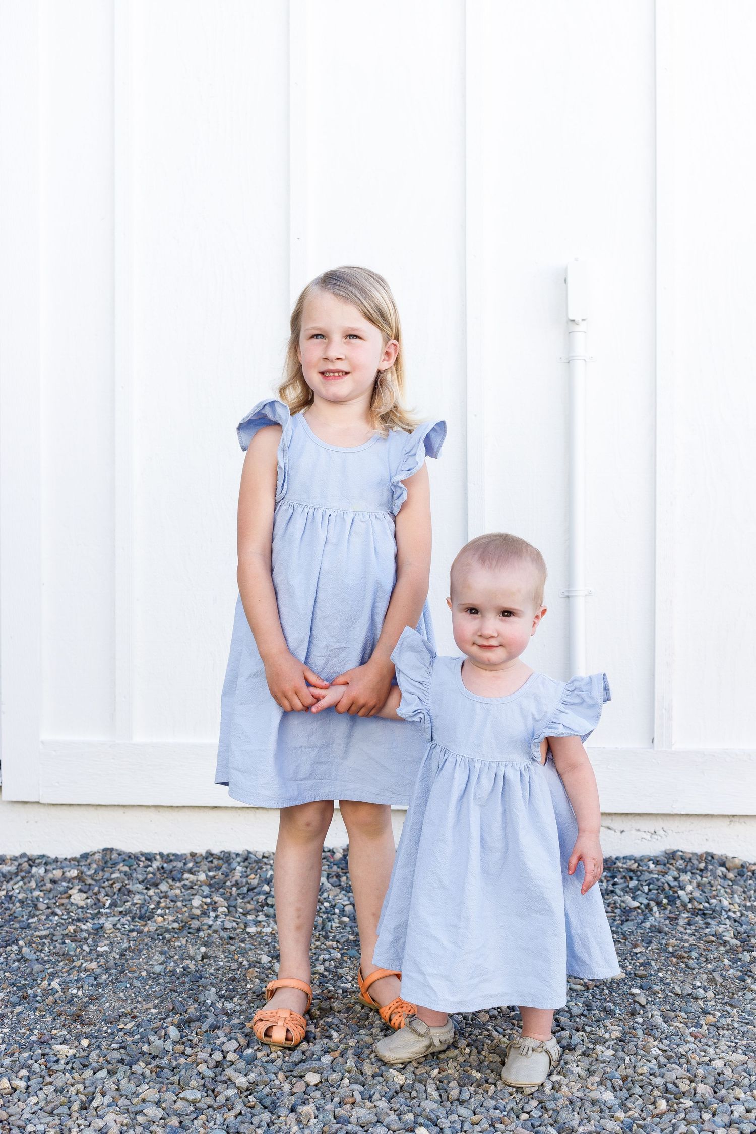 Three children in matching light blue dresses pose on gravel ground against a white wall background.
