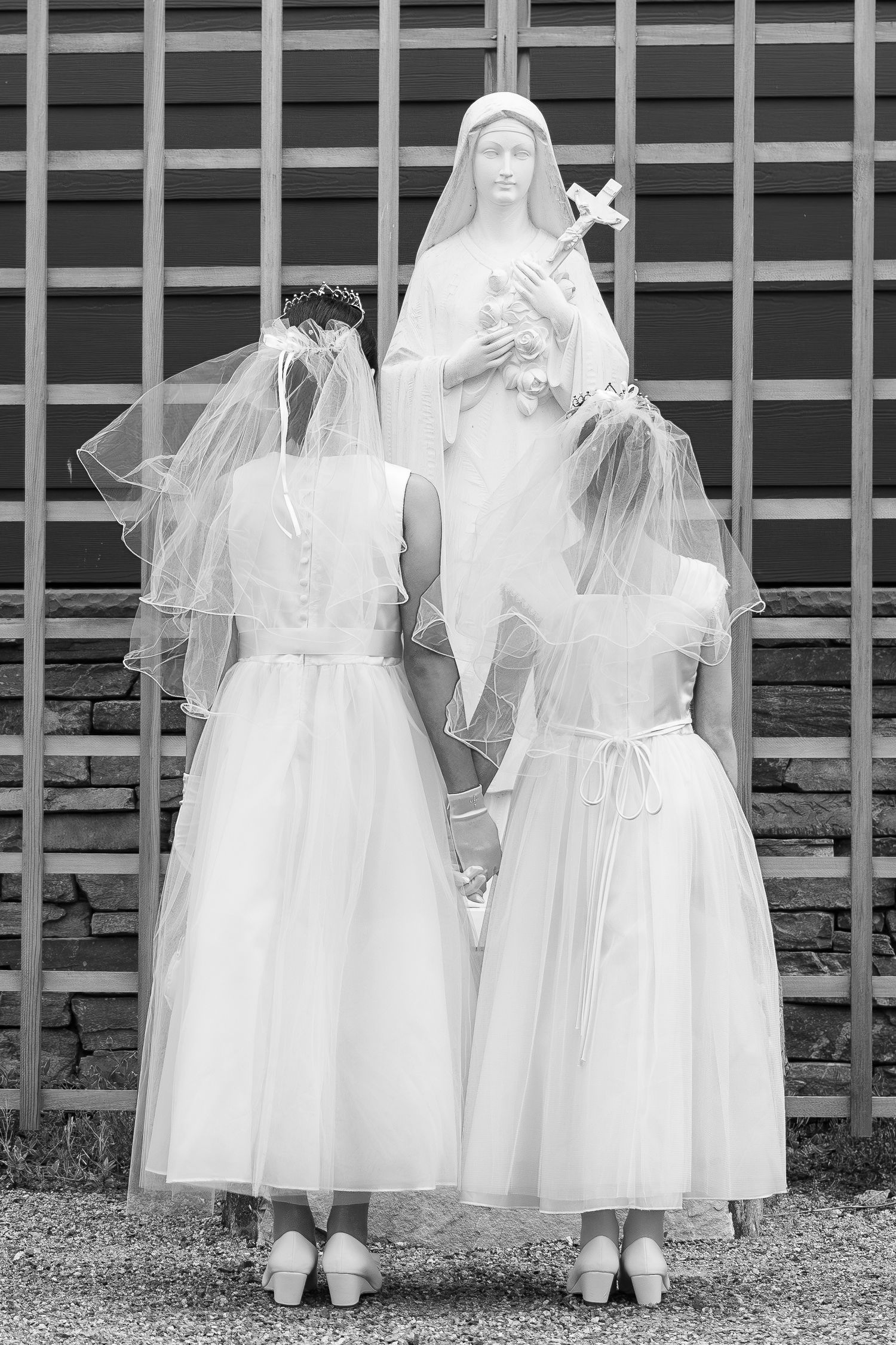 Two figures in white dresses and veils stand before a statue of religious figure holding a lily in black and white.