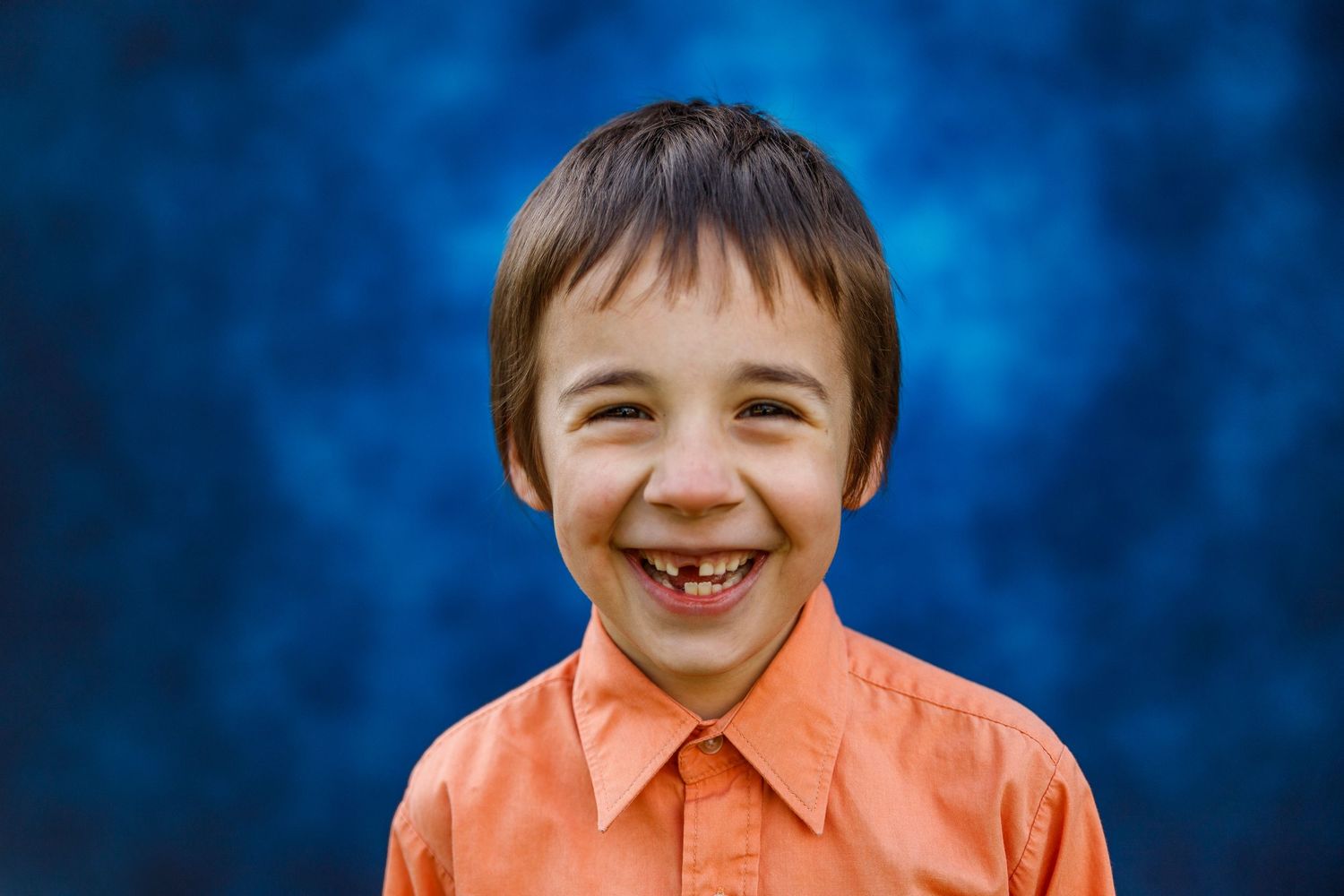 A young child in a bright orange collared shirt smiles warmly against a vibrant blue backdrop.