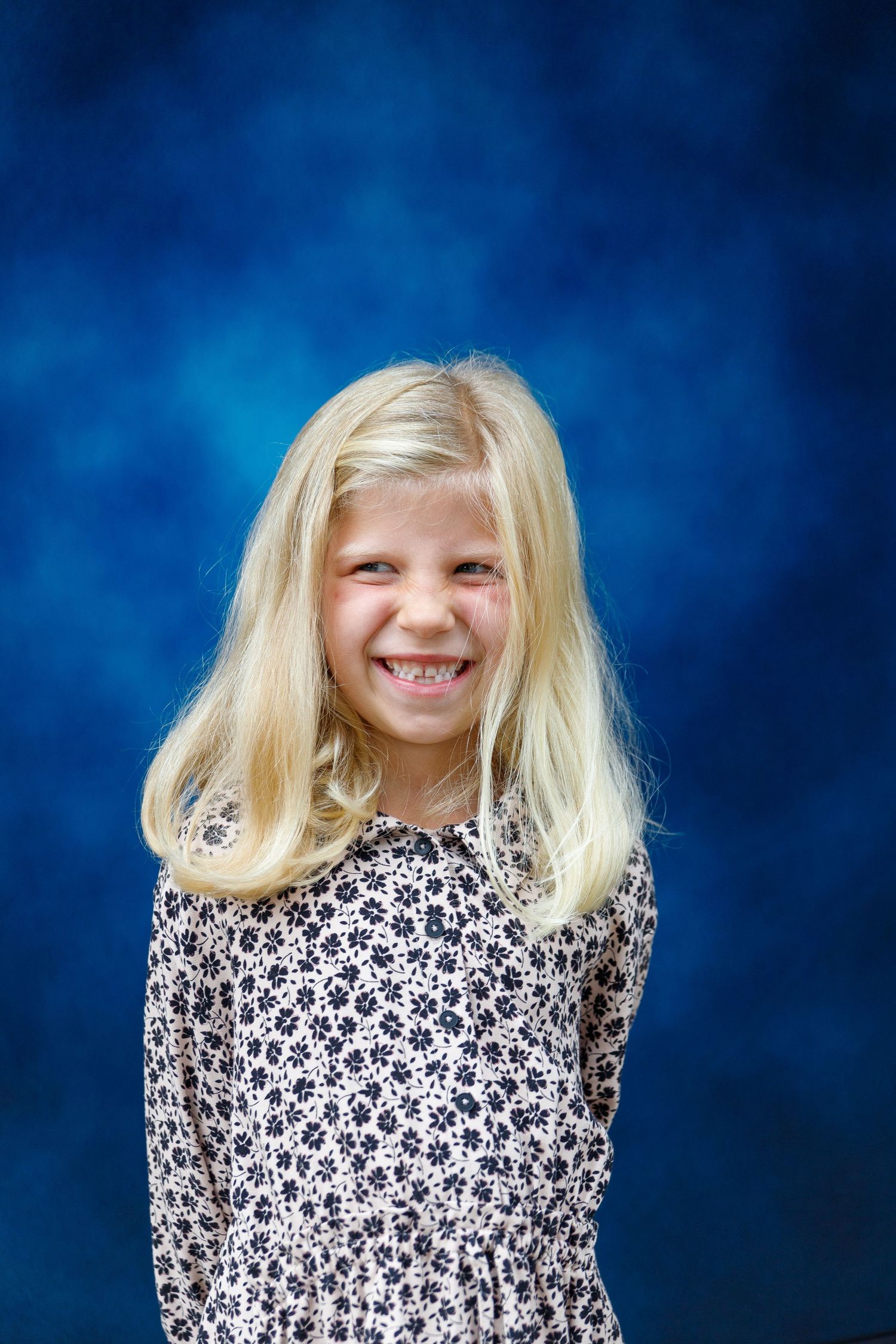School portrait with floral print dress against blue background shows bright smile and blonde hair.
