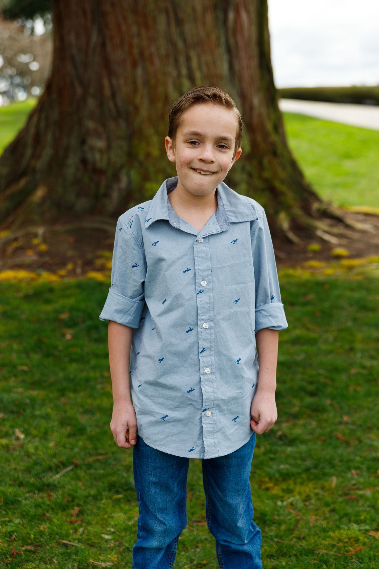 A youth in a light blue button-down shirt stands in front of a large tree trunk in a park setting.