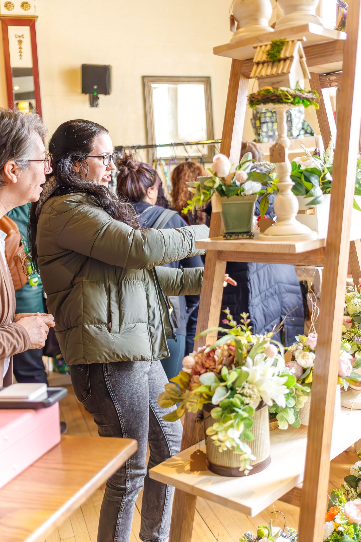 People examine potted plants and floral arrangements displayed on wooden shelves in a bright, cozy shop interior.