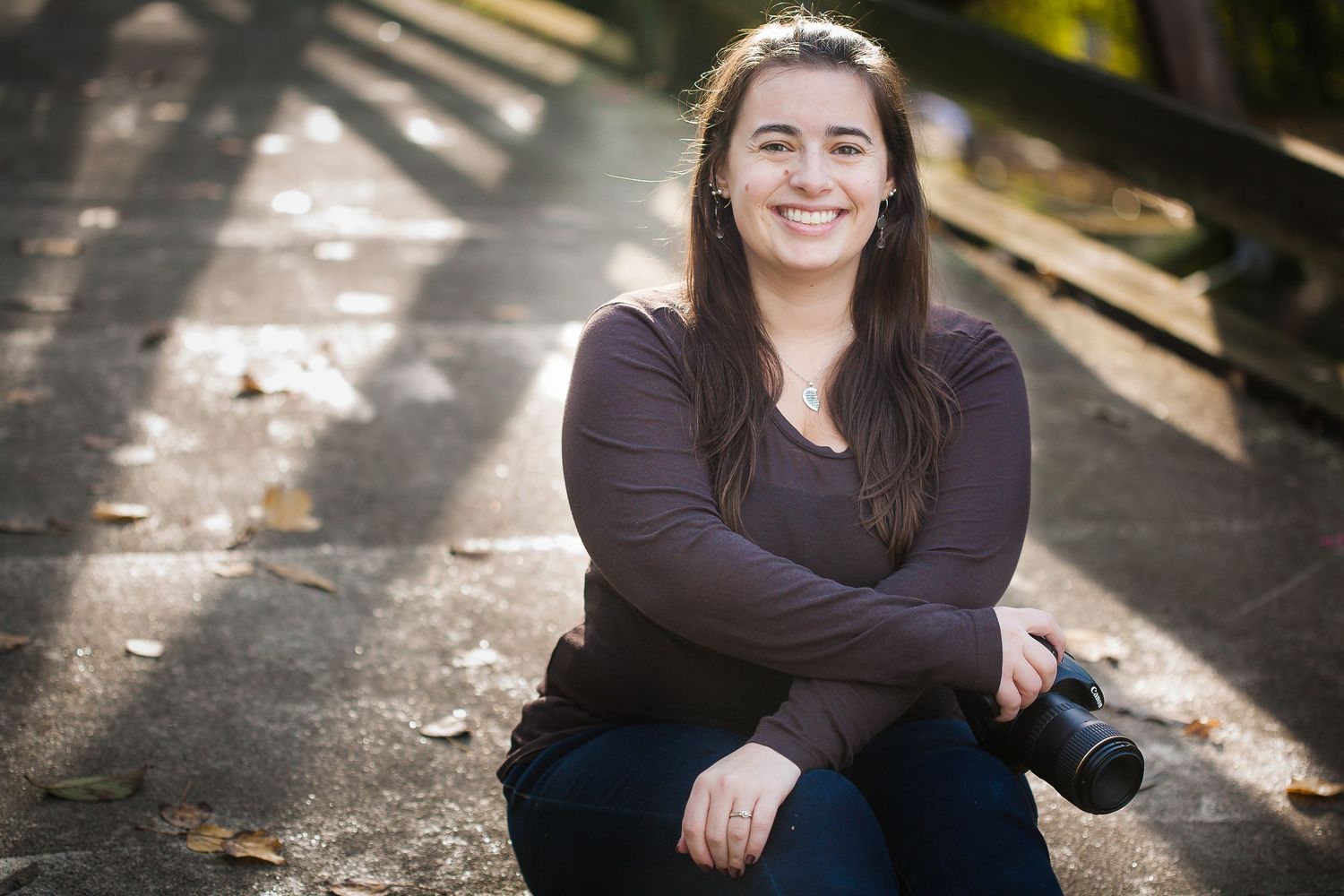 Person in dark sweater sits relaxed outdoors in dappled sunlight with a warm smile.