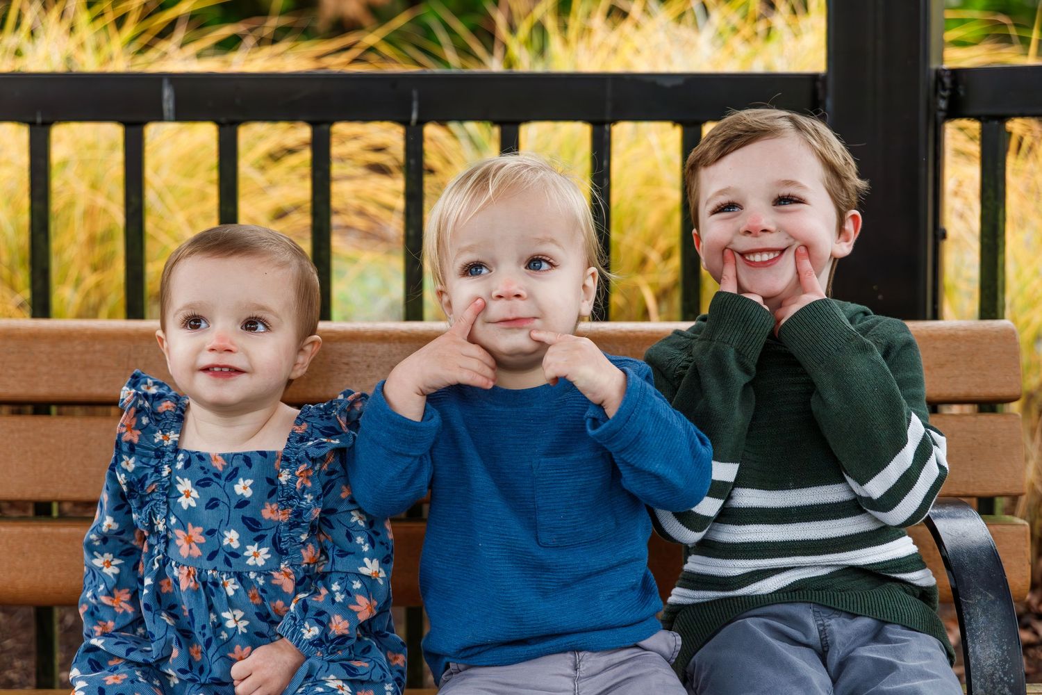 Three young children sitting together on a wooden bench and making silly expressions.