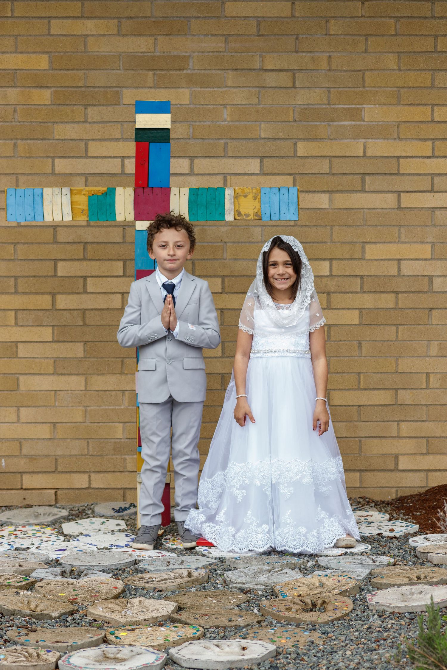 Two children in formal attire stand in front of colorful cross on brick wall for First Communion photo.