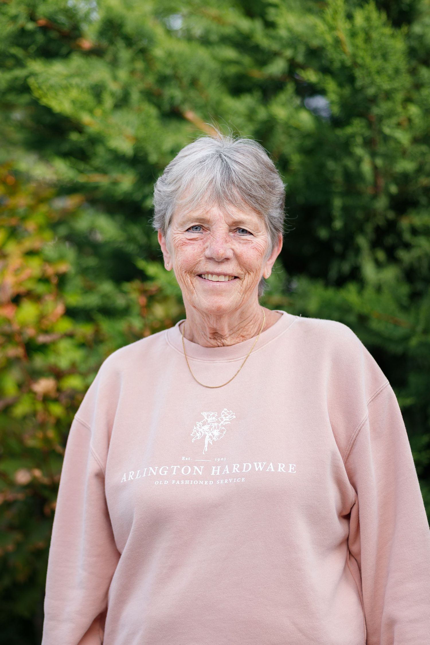 A person wearing a light pink sweatshirt smiles while standing outdoors against a green foliage background.