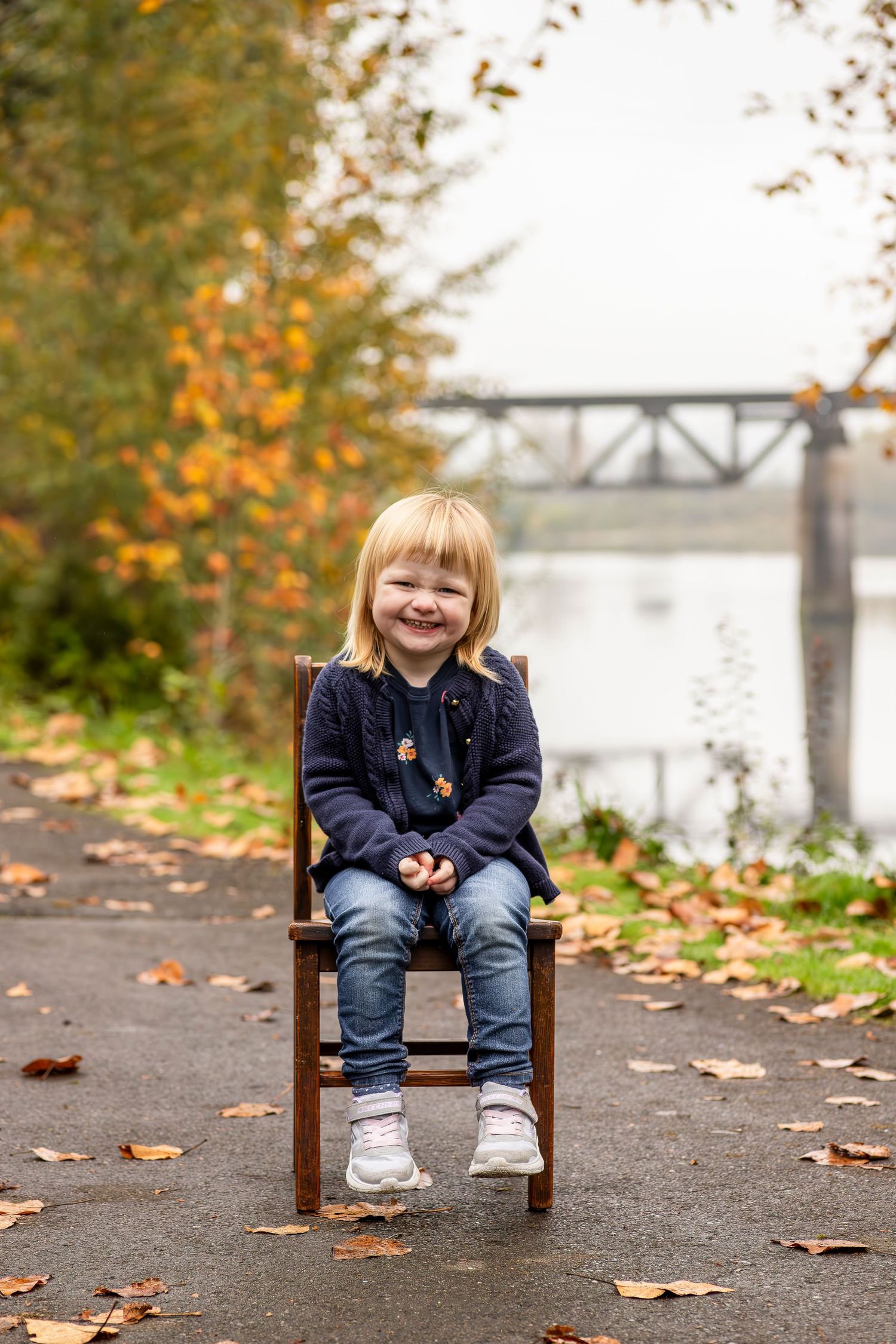A young child sits on a wooden chair along a riverside path with autumn leaves and a bridge in the background.