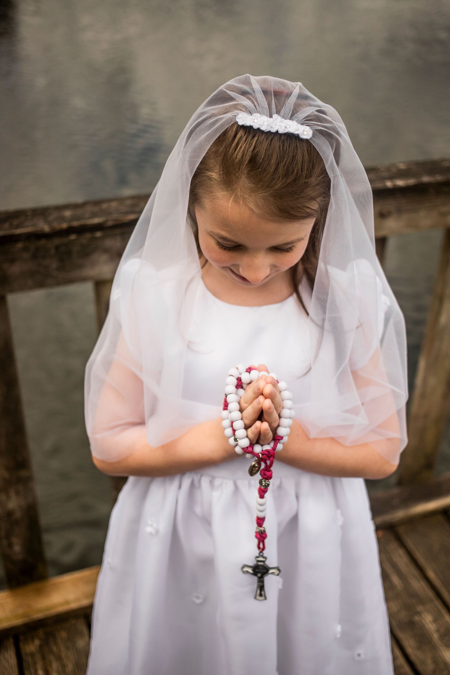 A figure in a white communion dress and veil holding prayer beads while looking down in prayer.