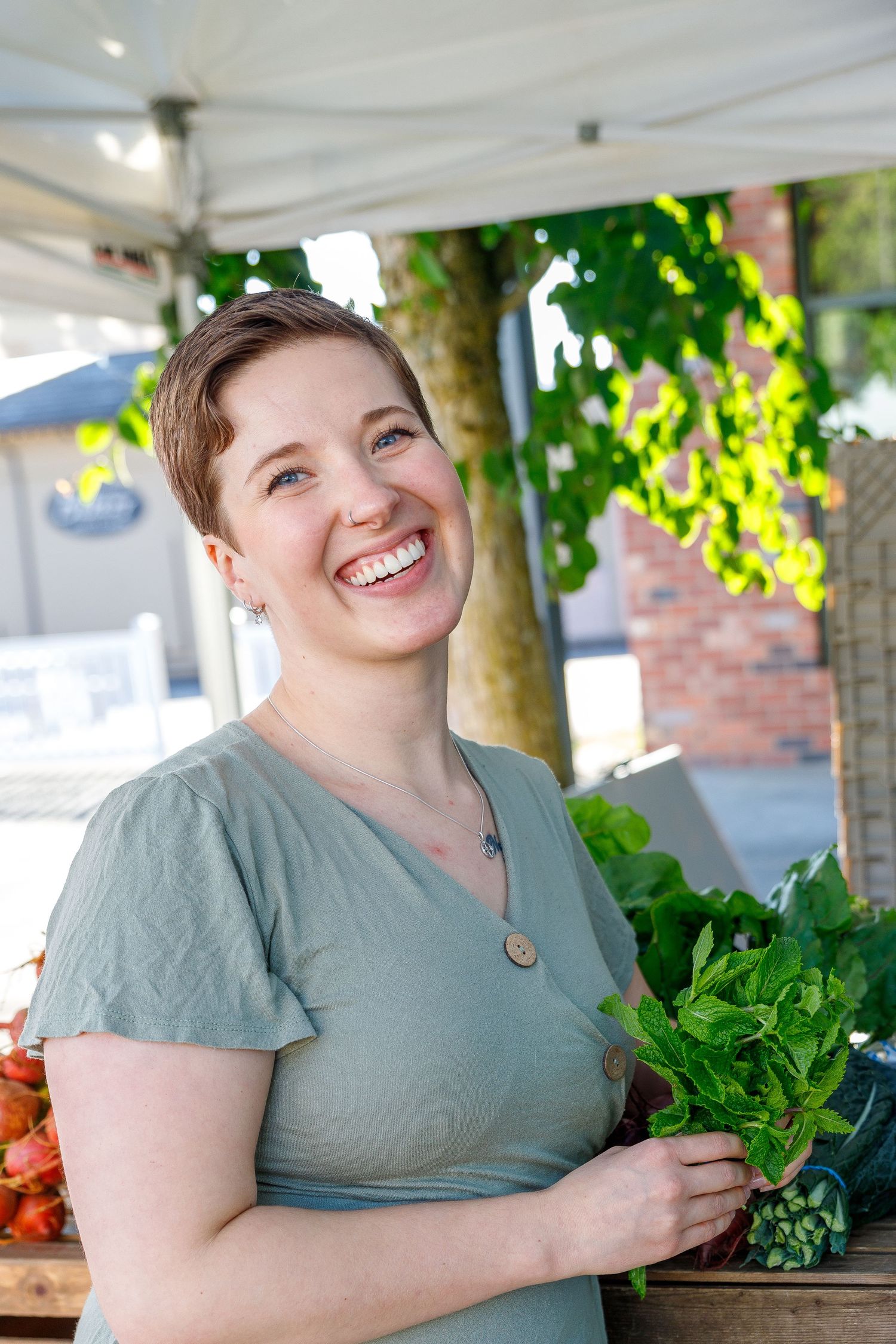 Person in gray shirt sits at outdoor table with fresh leafy vegetables and produce at farmers market.