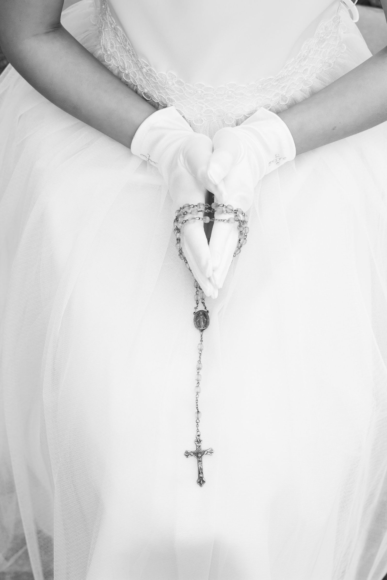 Close-up of gloved hands holding ornate silver rosary against white dress fabric.