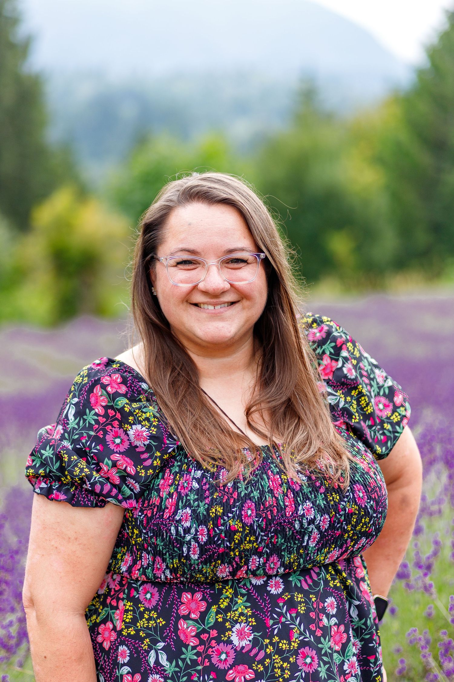 A smiling woman in a floral dress poses in a sunny lavender field with mountains and evergreen trees in the background.