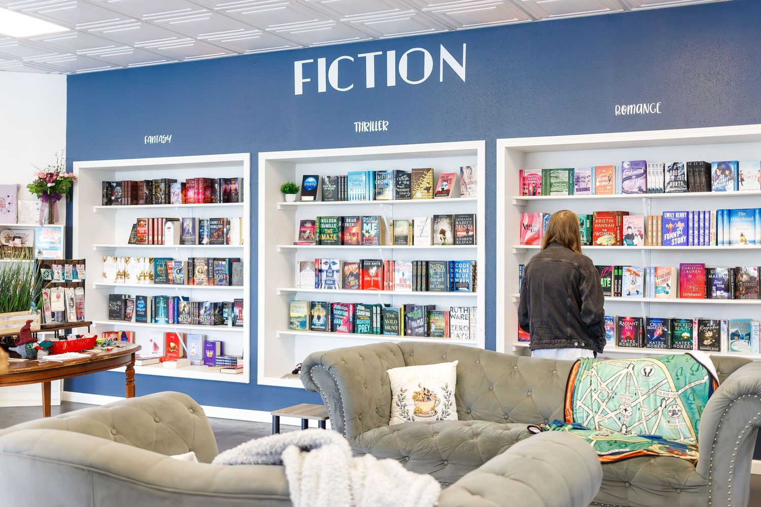 Cozy bookstore interior showing fiction section with white bookshelves and comfortable gray seating area.