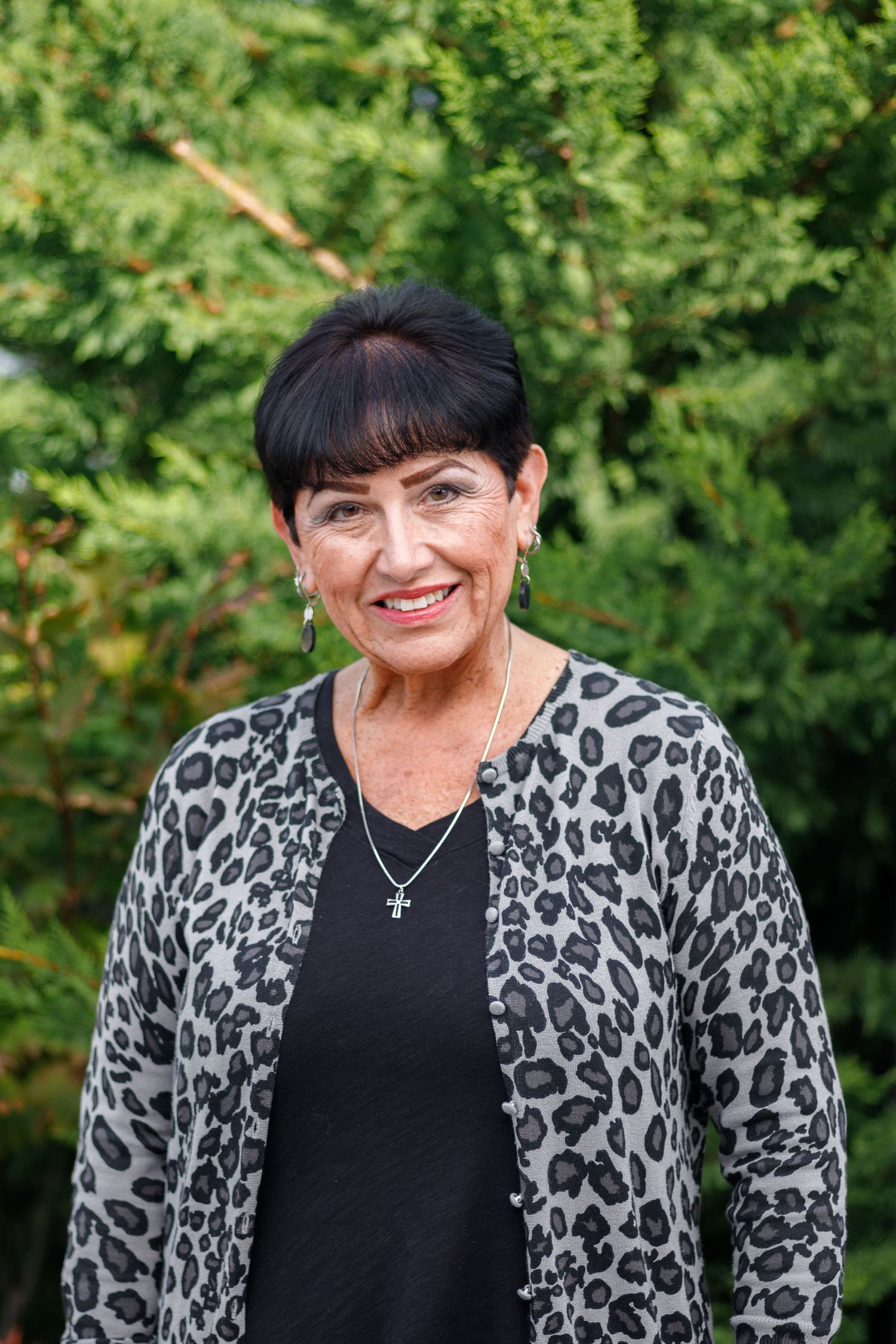 Person wearing black top and leopard print cardigan poses outdoors against green foliage background.