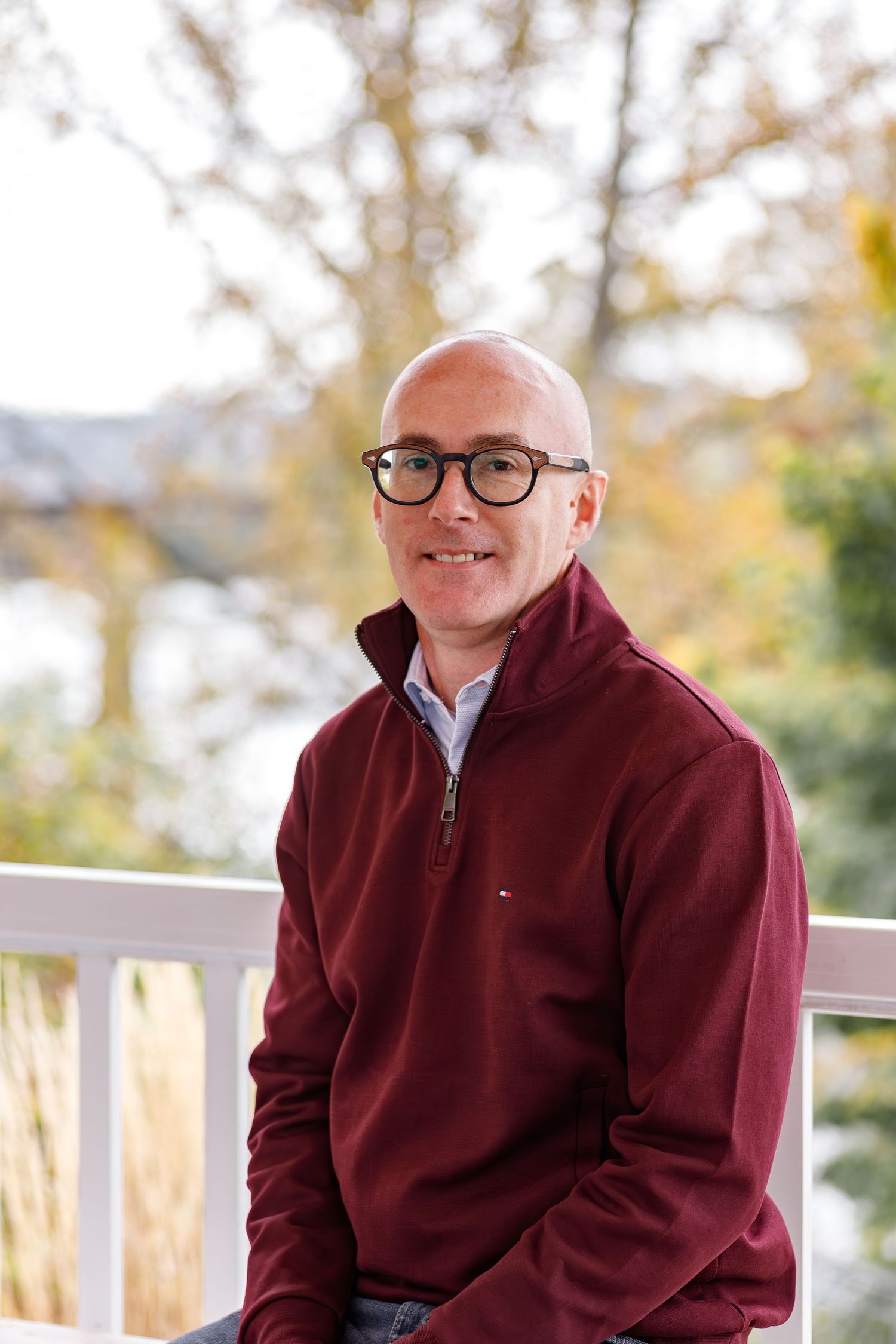 A person wearing glasses and a burgundy fleece sits outdoors on a deck during autumn.