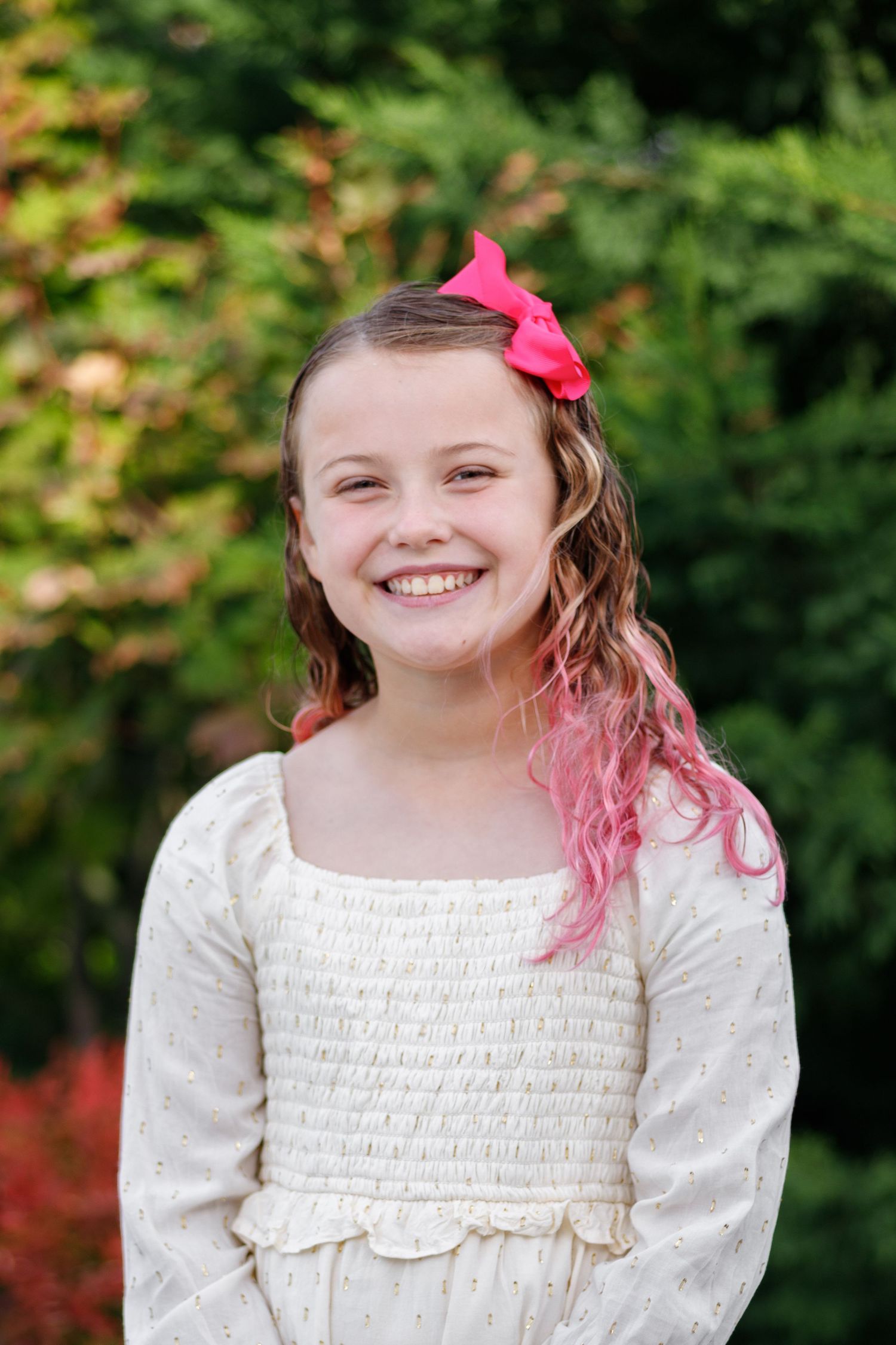 A person in a white smocked dress with pink hair and bow poses outdoors against a green background.