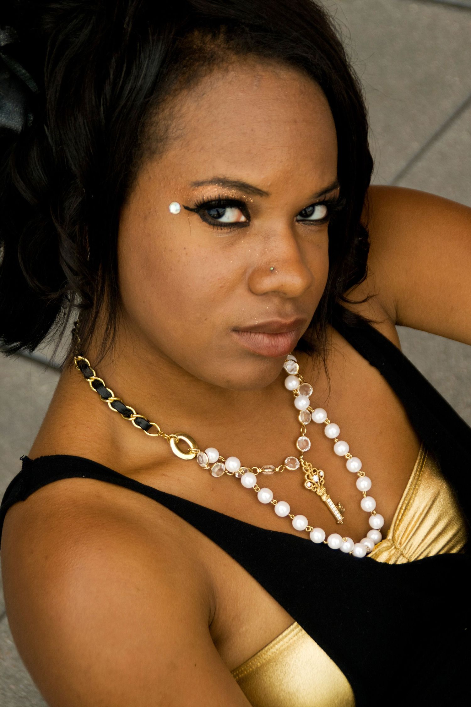 Portrait featuring dramatic eye makeup, black and gold top, and layered pearl and chain necklace accessories.