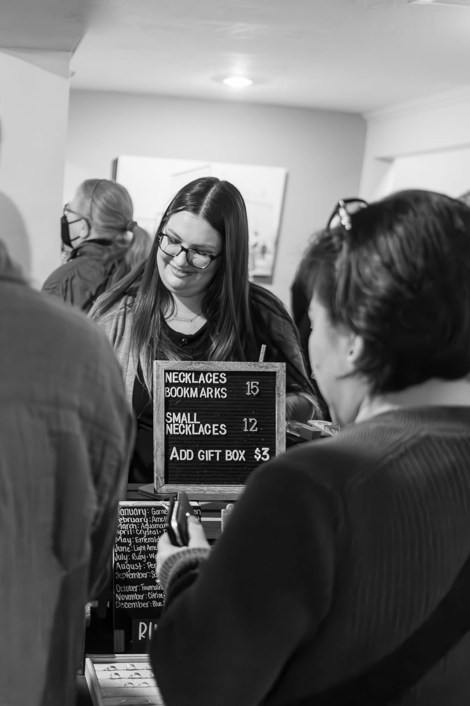 A black and white photo showing a person wearing glasses behind a counter with a menu board displaying necklace prices.