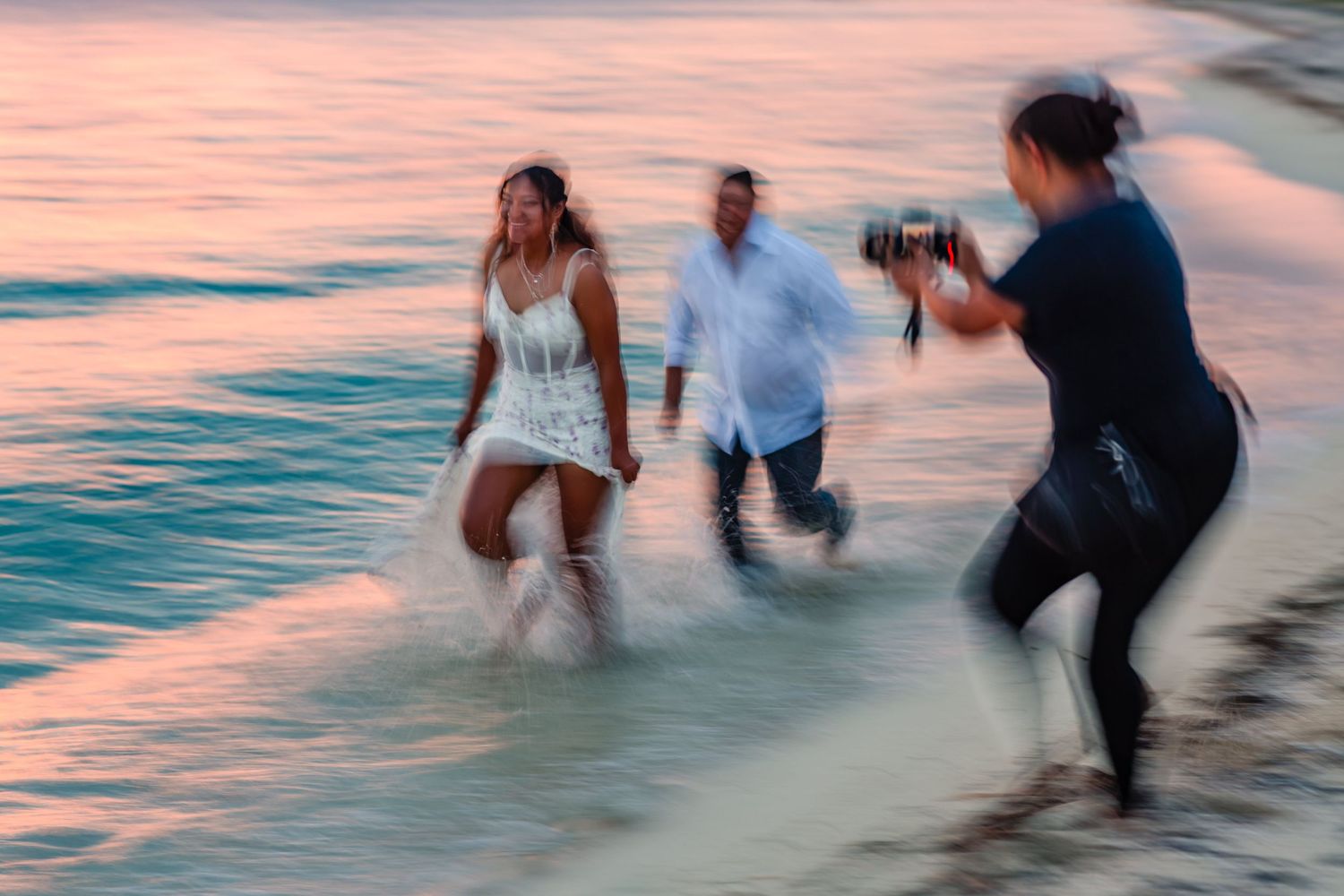Couple in white attire wading through ocean waves at sunset while photographer Renata Corichi captures the moment on beach in Cozumel