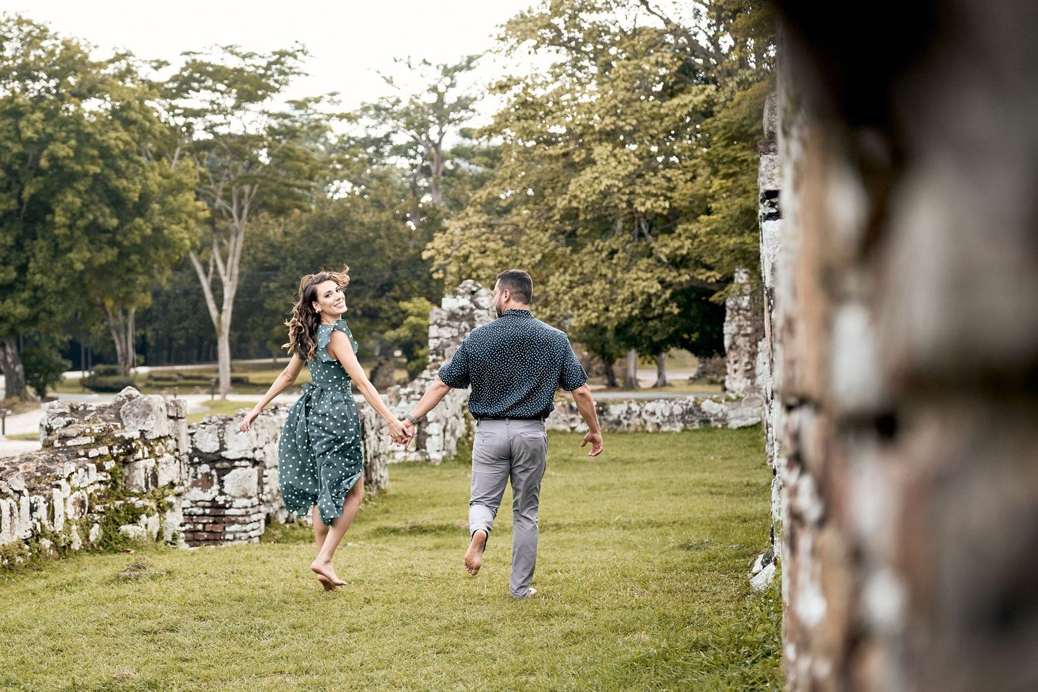 A couple runs joyfully through a grassy field with trees in the background during a photo session.