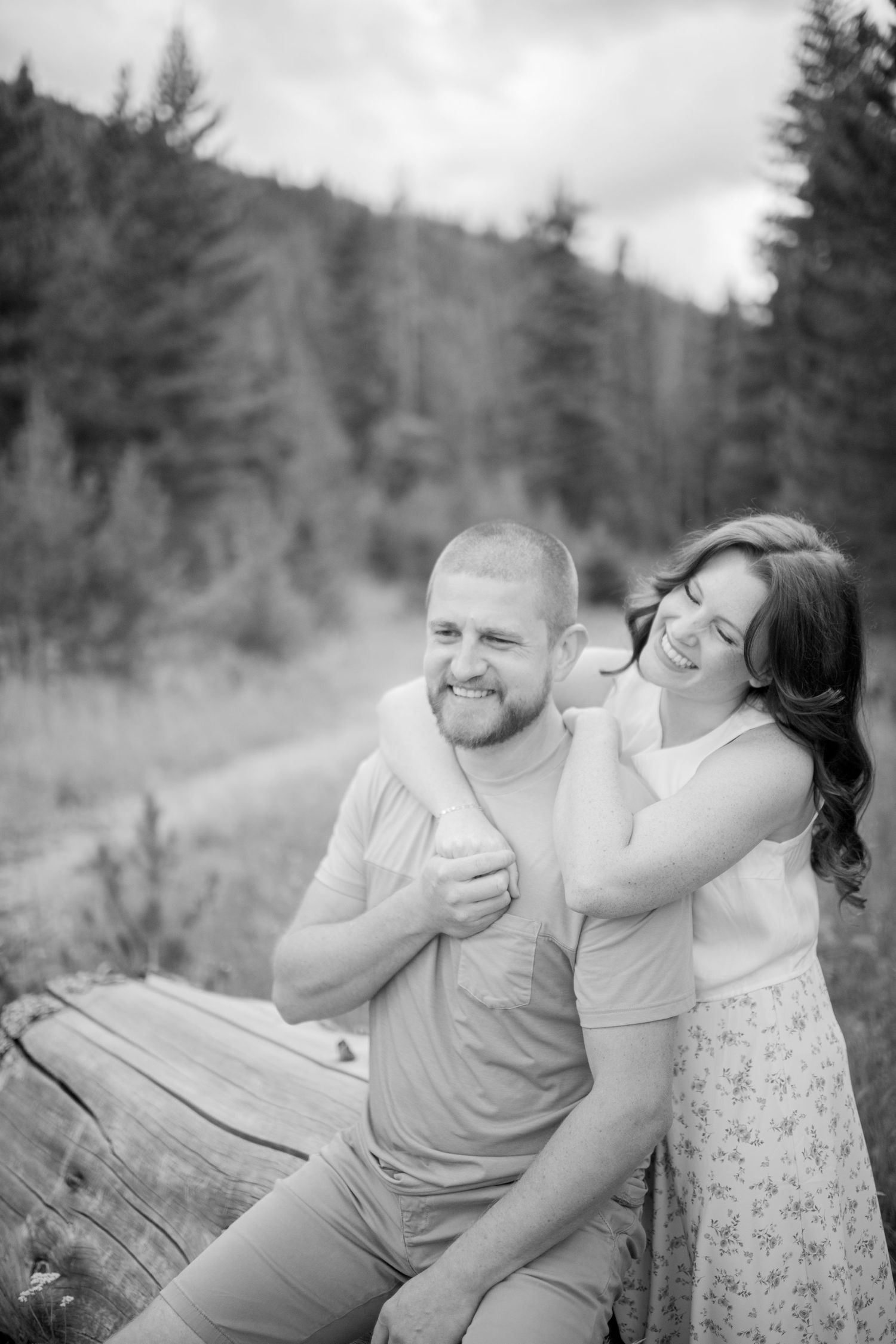 A couple shares playful laughter while sitting together outdoors in a forest setting with evergreen trees in the background.