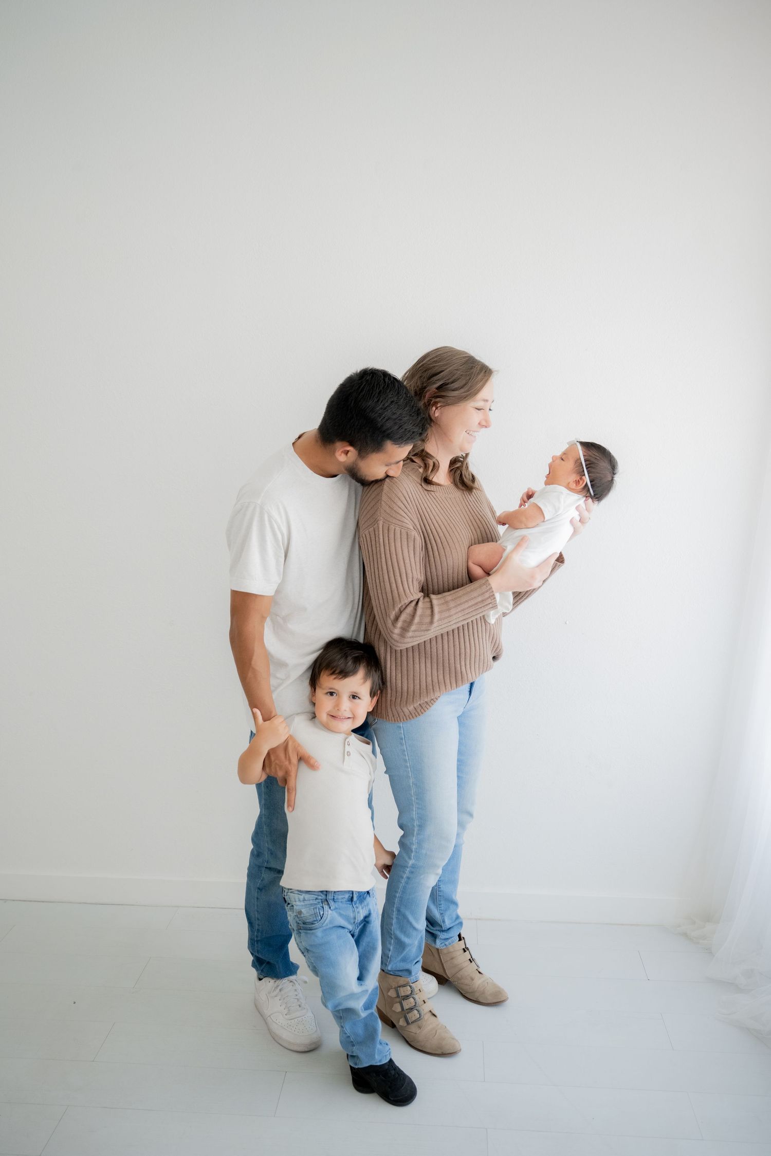 Family portrait with parents and two children standing together against a white wall in casual attire.