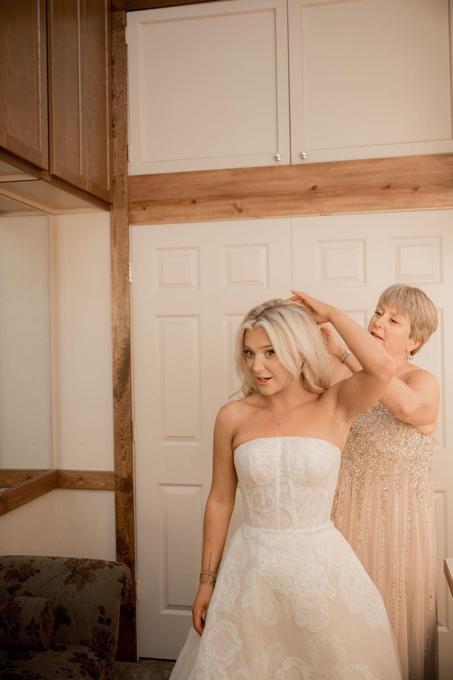 A bride in a strapless white lace wedding gown gets her hair styled in a rustic room with wooden beams.