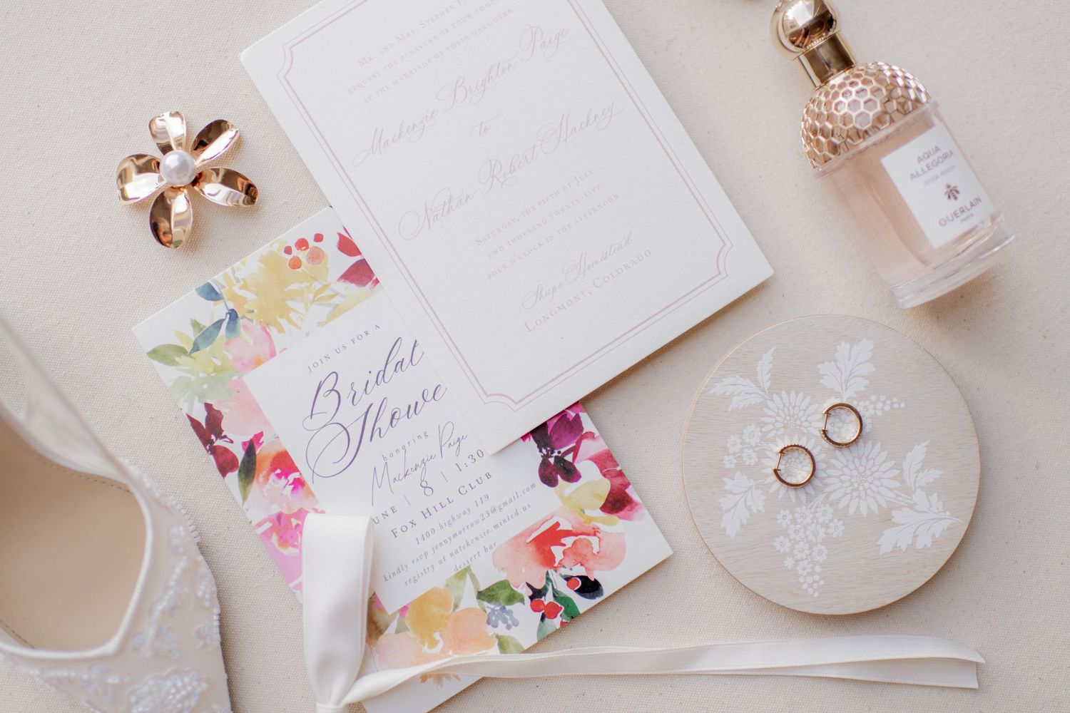 Wedding details flatlay with invitation cards, perfume bottle, rings, and delicate floral designs in soft pastel colors.