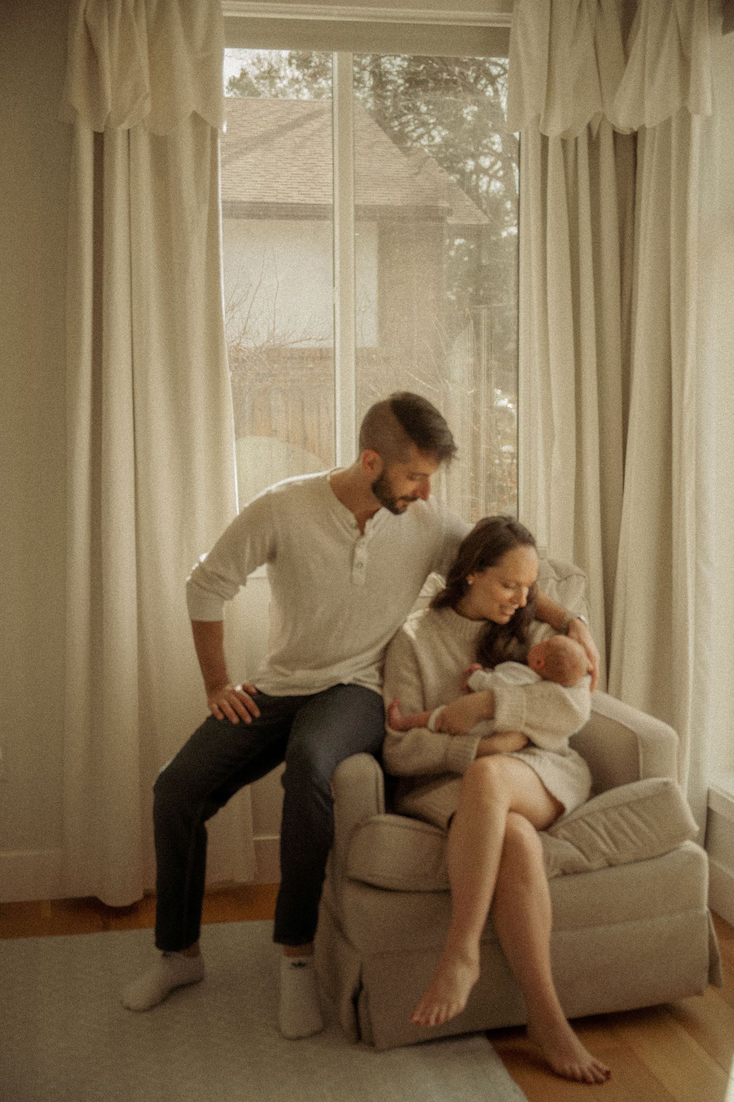 A family moment in a sunlit room with cream curtains as they sit together in a cozy armchair by the window.