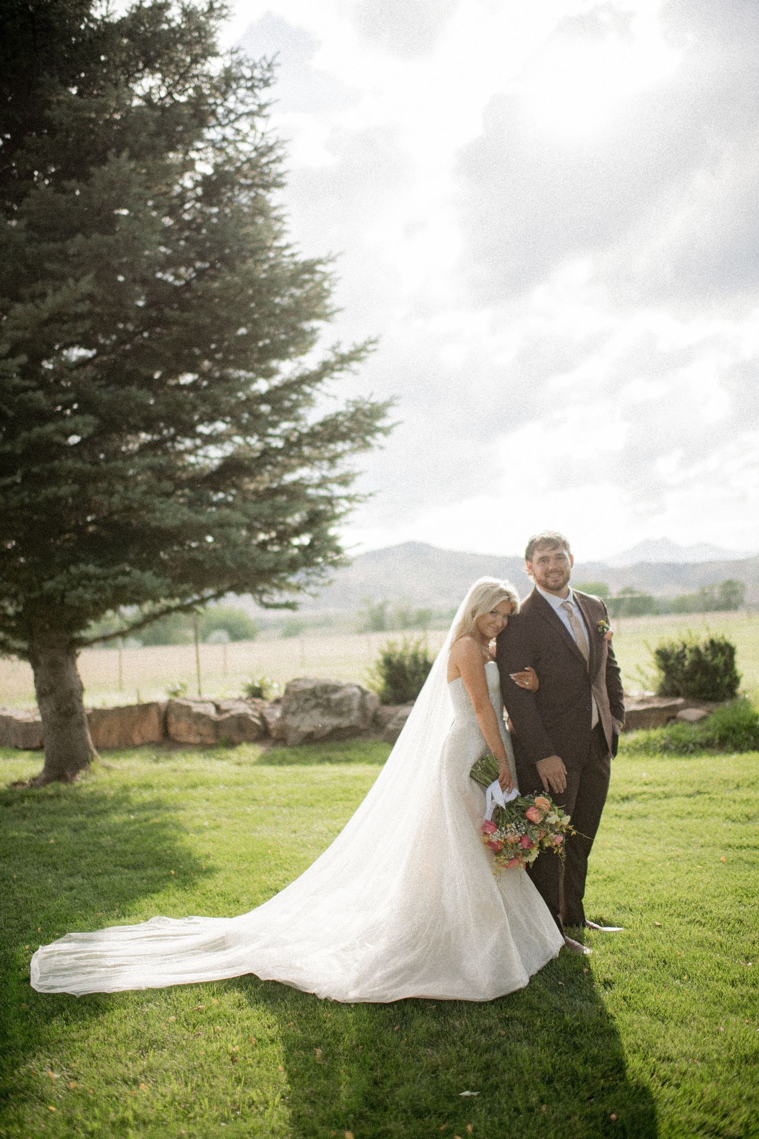 A romantic wedding couple poses together on a grassy hillside with evergreen trees and mountains in the background.