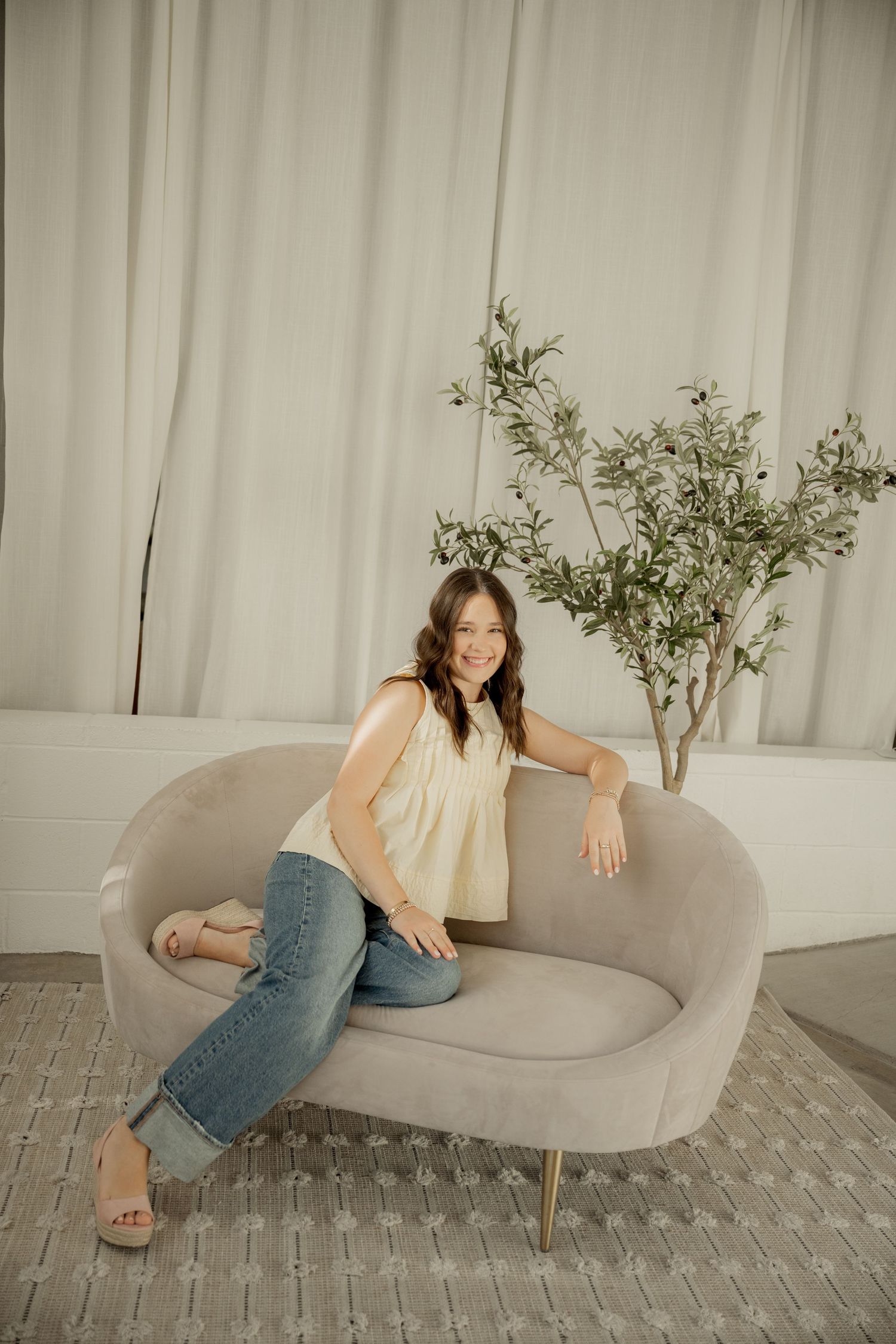 Woman in jeans and yellow top lounges on modern gray chaise sofa with potted olive tree in background.
